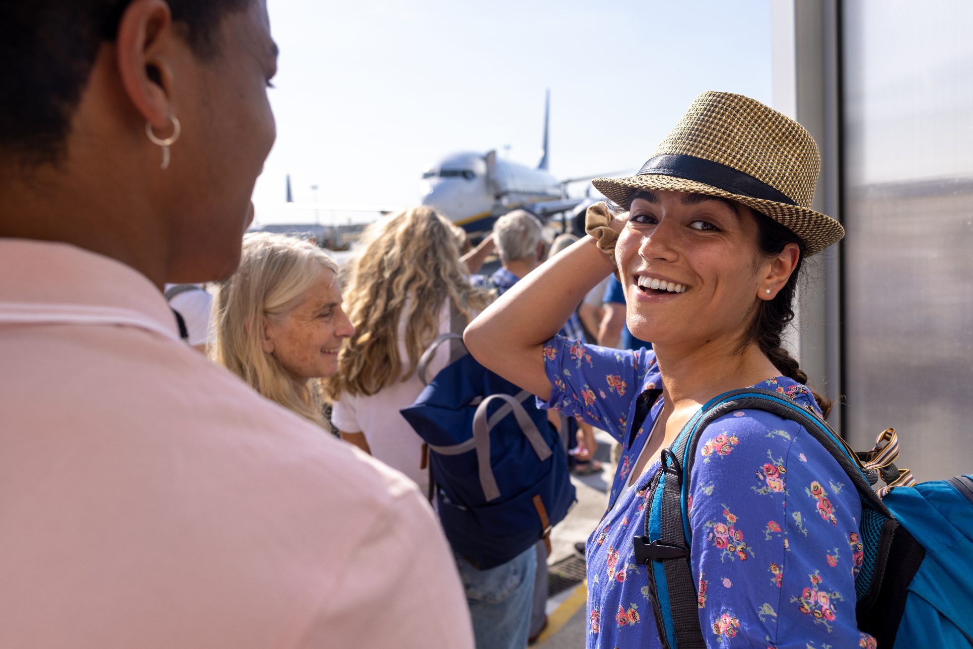 An over the shoulder shot of a young man and woman standing in a queue outdoors on an airport runway in Toulouse, France waiting to get on their flight. The young woman is looking at the camera and smiling while wearing a trilby hat.