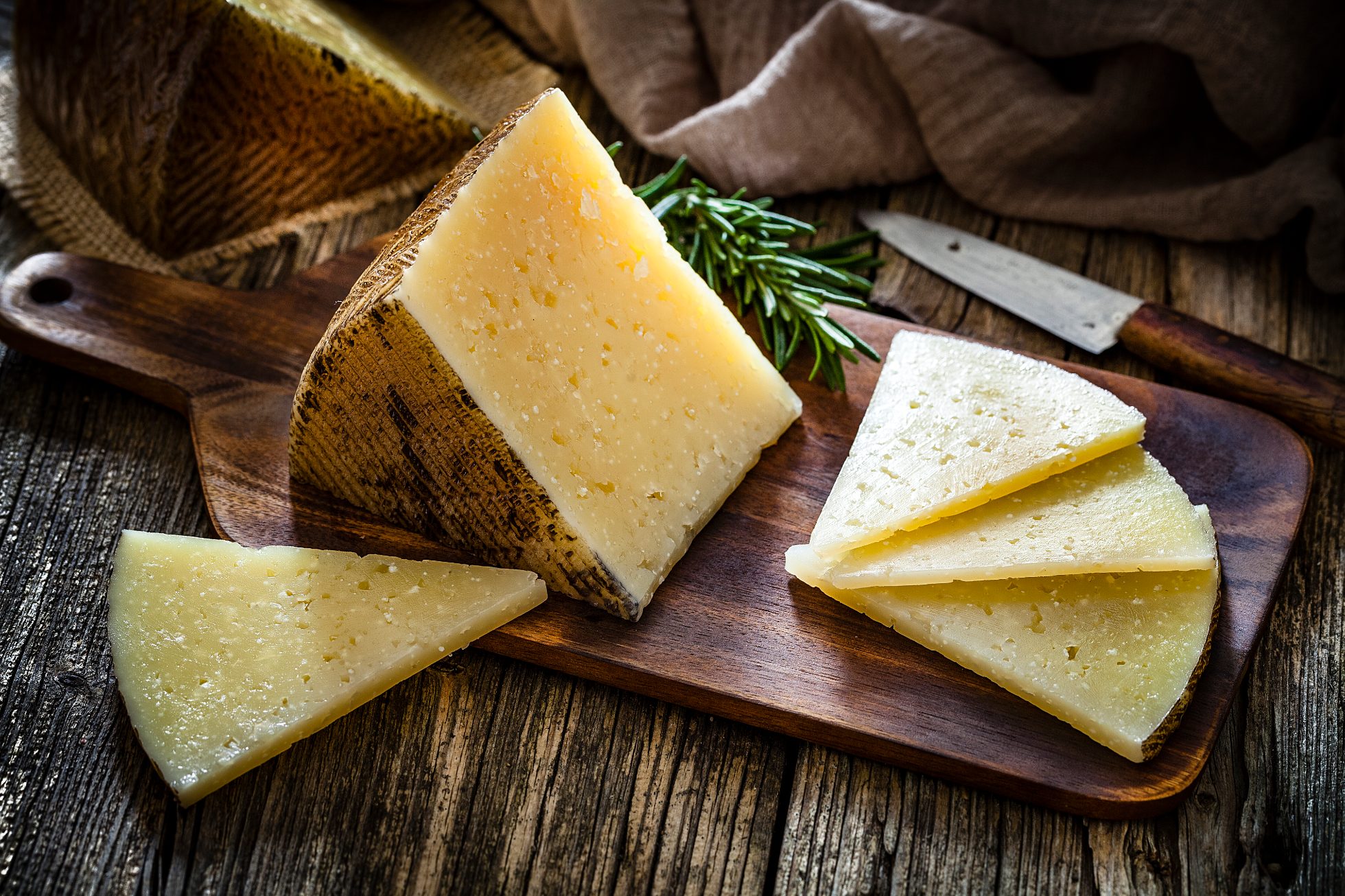 Hiszpańskie potrawy: kawałki i plasterki sera Manchego na desce do krojenia, sfotografowane na ciemnym, rustykalnym drewnianym stole. Za kawałkami sera leży stary nóż. Dominują kolory brązowy i żółty. Stonowane zdjęcie studyjne