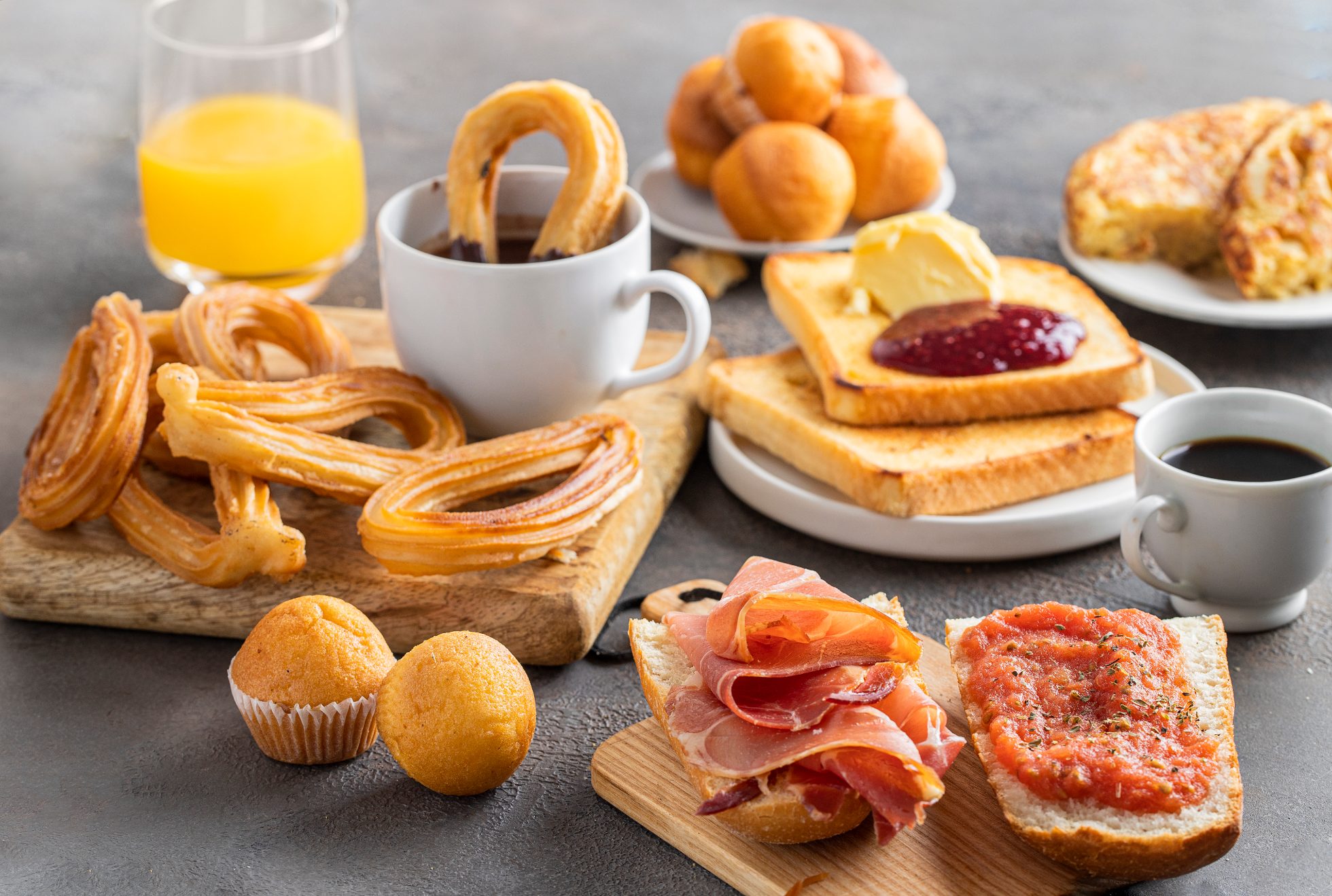 Różnorodne hiszpańskie śniadanie: tortilla, bruschetta z szynką jamón i pomidorami, churros z czekoladą, magdalena (hiszpańska babeczka), chrupiący tost francuski z masłem i dżemem, kawa i sok pomarańczowy.