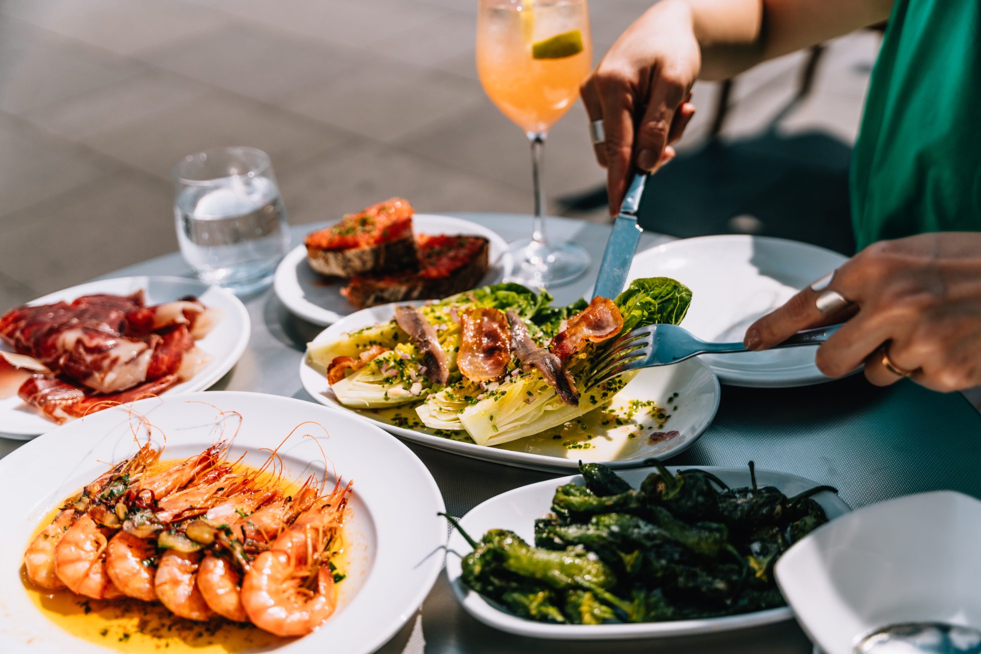 Zbliżenie na młodą kobietę jedzącą hiszpańskie potrawy w restauracji na świeżym powietrzu. Na stole podano sałatkę z sałaty rzymskiej, grillowane krewetki, plasterki szynki iberyjskiej, chleb z pomidorem oraz papryczki Padrón. Hiszpańska kuchnia i kultura.