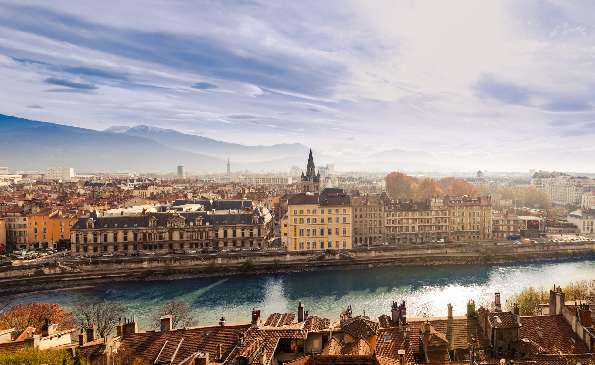 Panorama Grenoble w mglisty dzień. W tle pasmo Alp, a zza chmur wyłania się słońce. Grenoble, Francja.