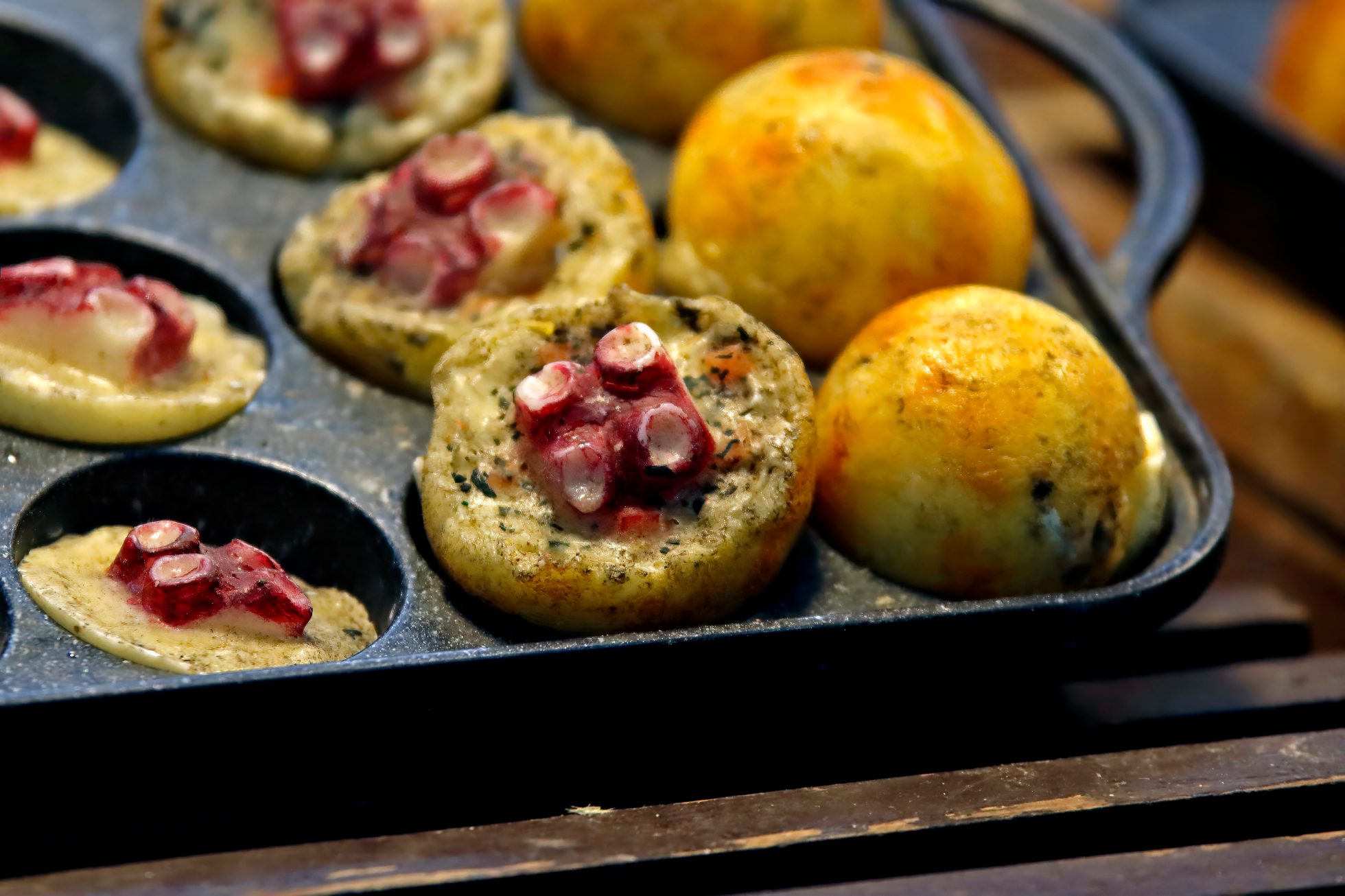 Zbliżenie na świeże japońskie takoyaki smażone na czarnej żeliwnej patelni, ukazujące złocistobrązowe kuliste kształty i widoczne małe kawałki macki ośmiornicy – popularną azjatycką przekąskę uliczną