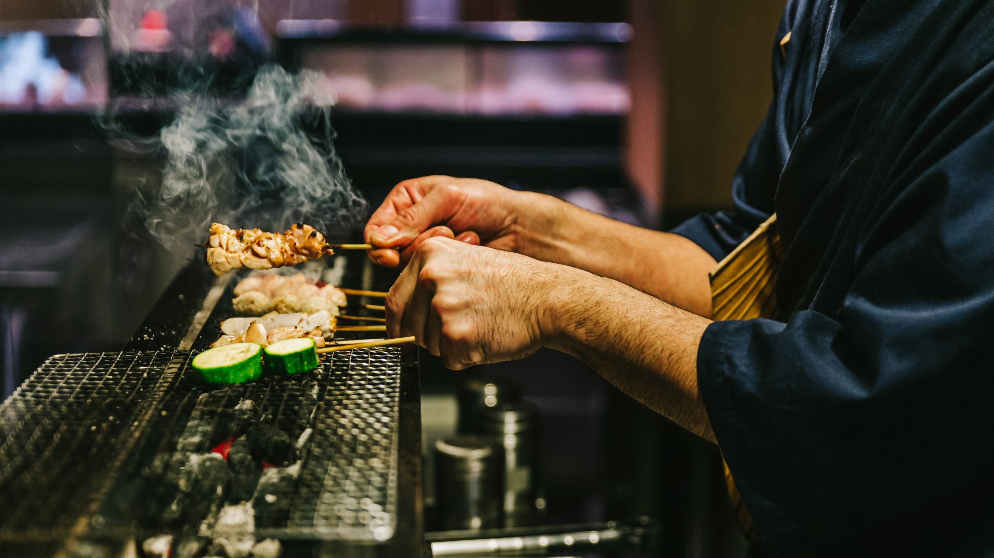 Ręce japońskiego szefa kuchni specjalizującego się w yakitori, który grilluje kurczaka marynowanego w imbirze, czosnku i sosie sojowym oraz ogórka, a wszystko to w gęstej chmurze dymu.