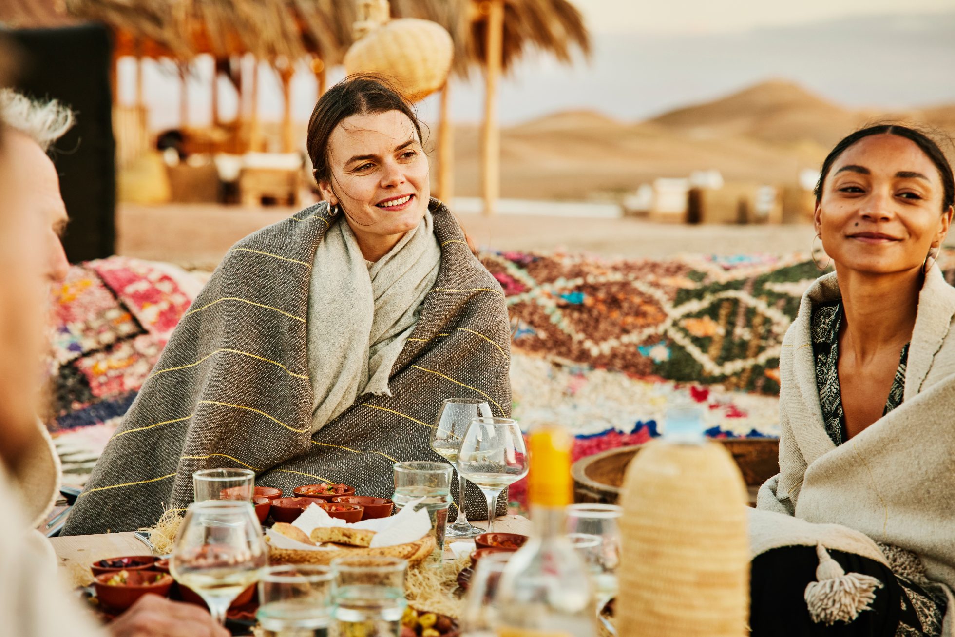 Medium shot of smiling female friends wrapped in warm blankets in discussion while sharing meal with family and friends during outdoor sunset dinner at luxury Moroccan desert camp