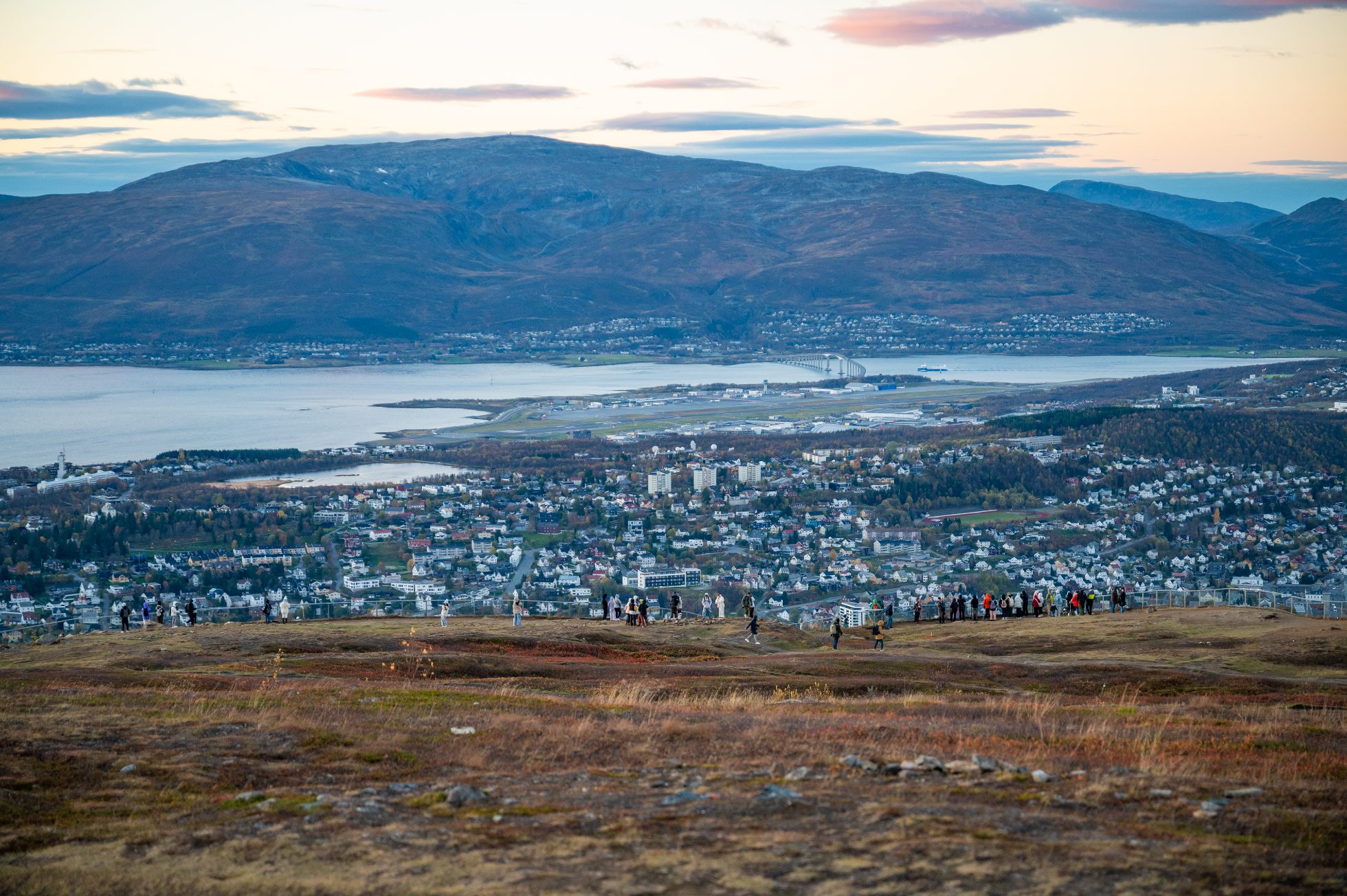 Widok ze Storsteinen w Tromsø w Norwegii – panoramiczny widok na miasto położone pomiędzy falistymi wzgórzami a rozległym akwenem, z ludźmi zgromadzonymi na szczycie wzgórza