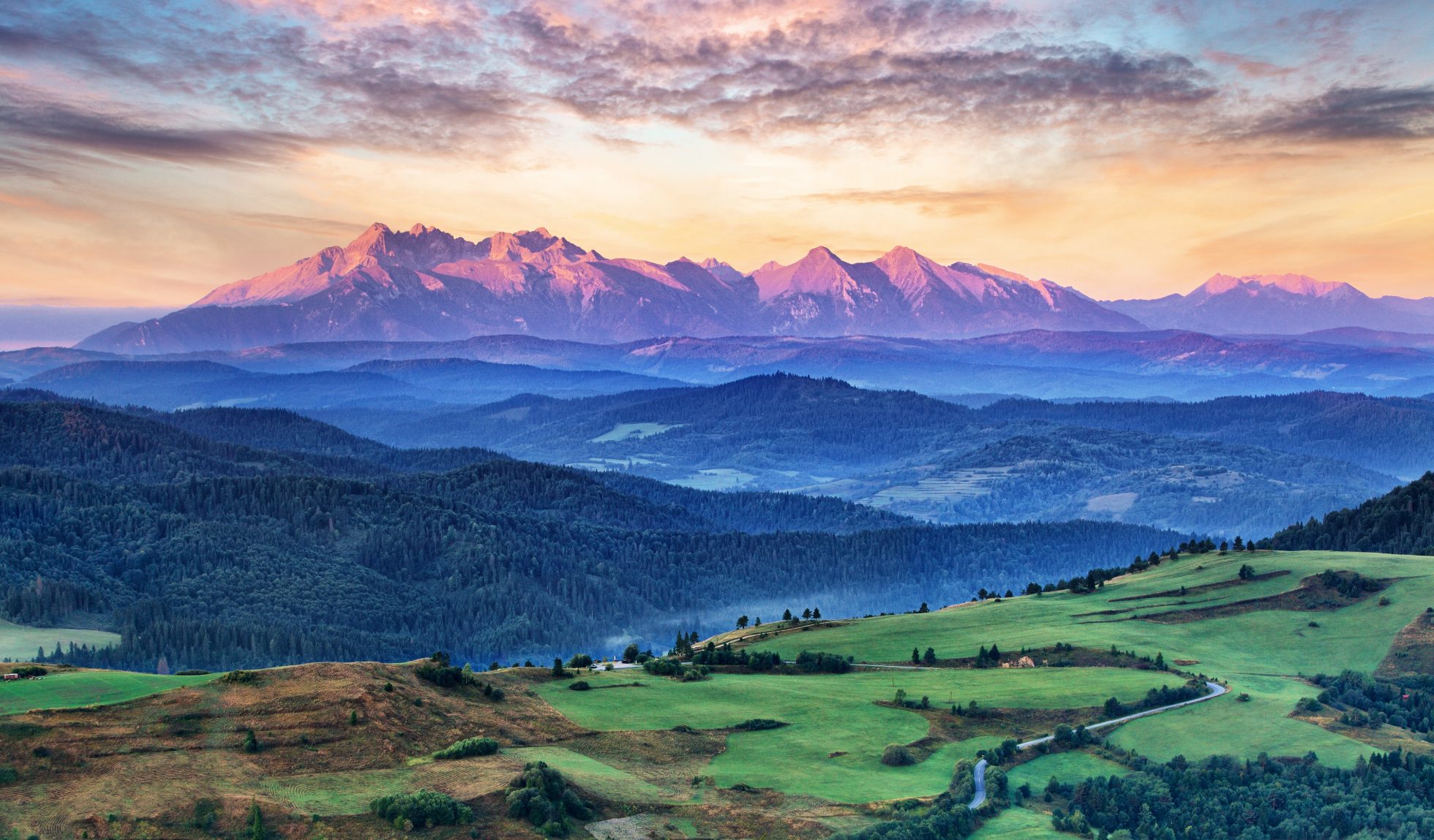 Górzysty krajobraz z widokiem na Tatry Polskie i Słowackie w oddali. Szlak turystyczny wśród zielonych łąk i szczytów gór.