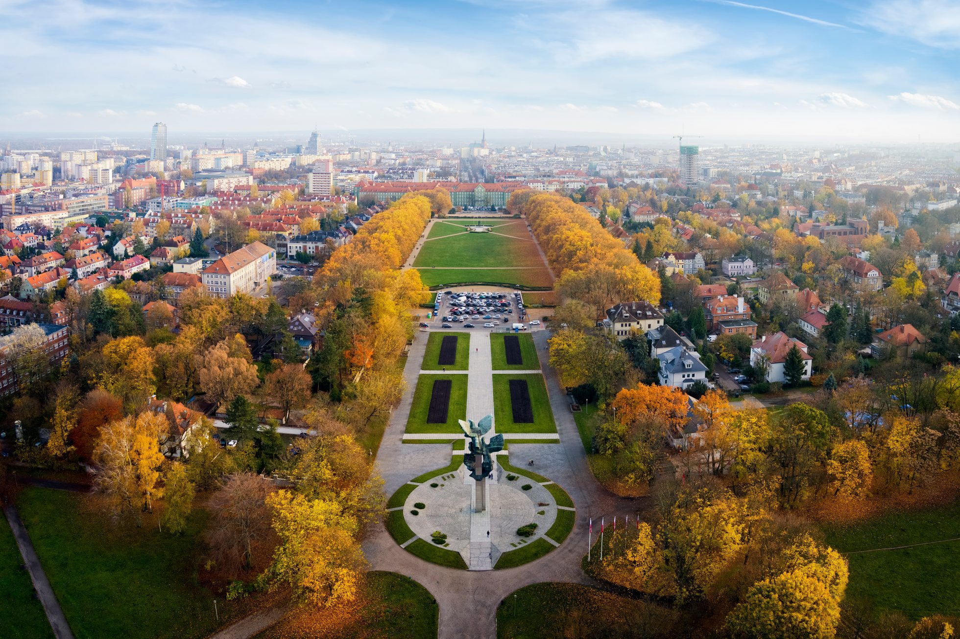 Park Kasprowicza w Szczecinie - zielona przestrzeń z drzewami, trawnikami i alejkami. W oddali widoczne ławki i fontanna.