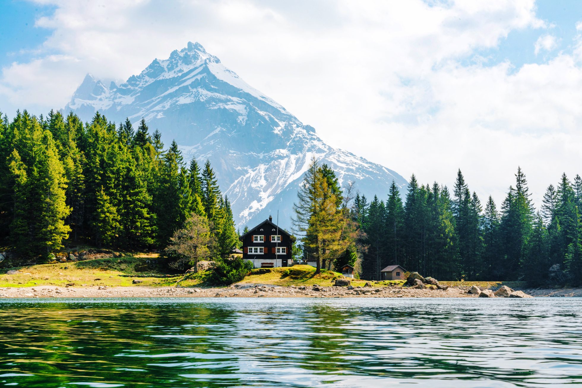 A cottage by the lake near the forest, with mountains in the background.