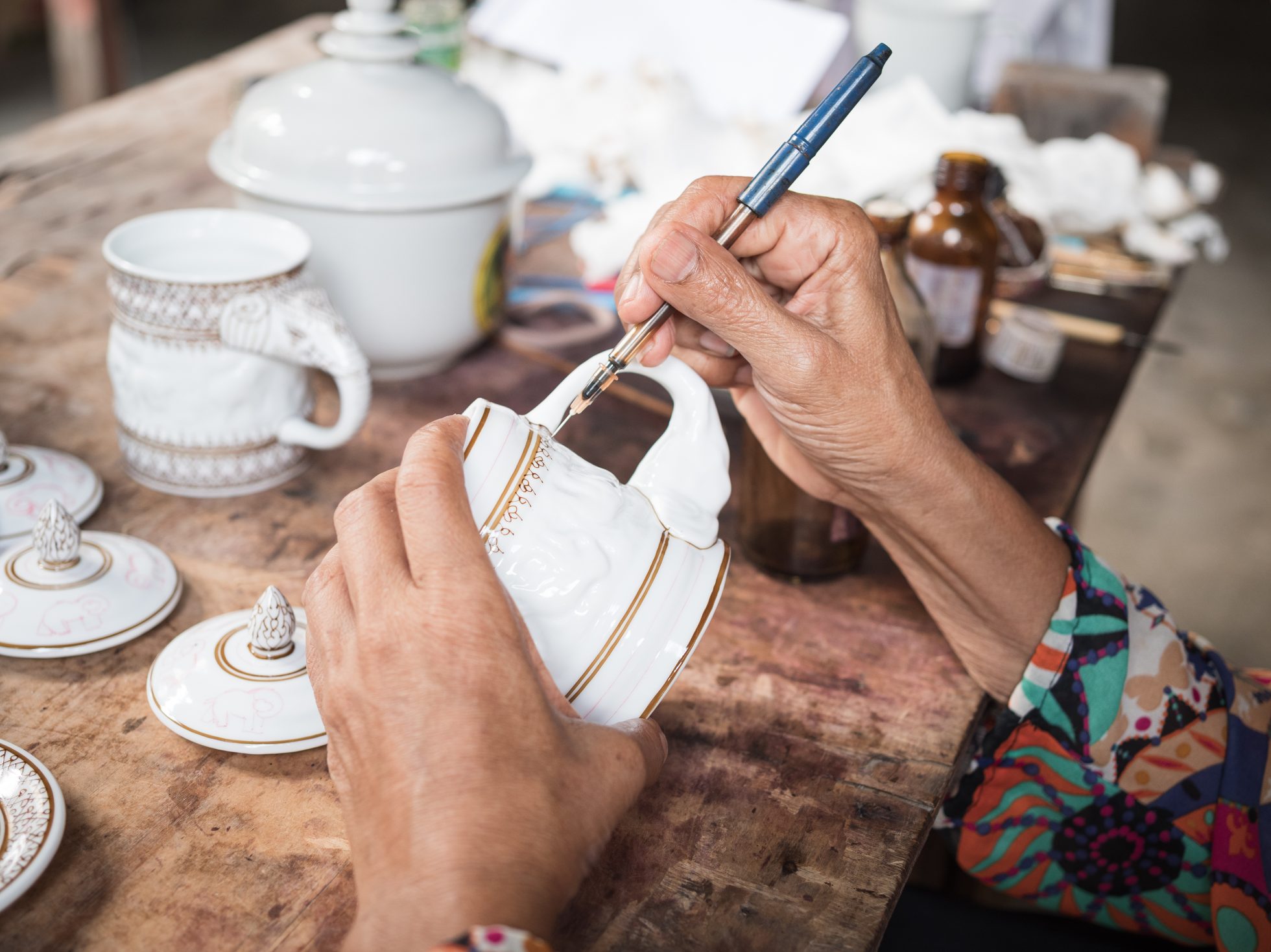 Ręce kobiety nakładającej złoty liść na biały ceramiczny kubek, a w tle na drewnianym stole widać inne ceramiczne przedmioty