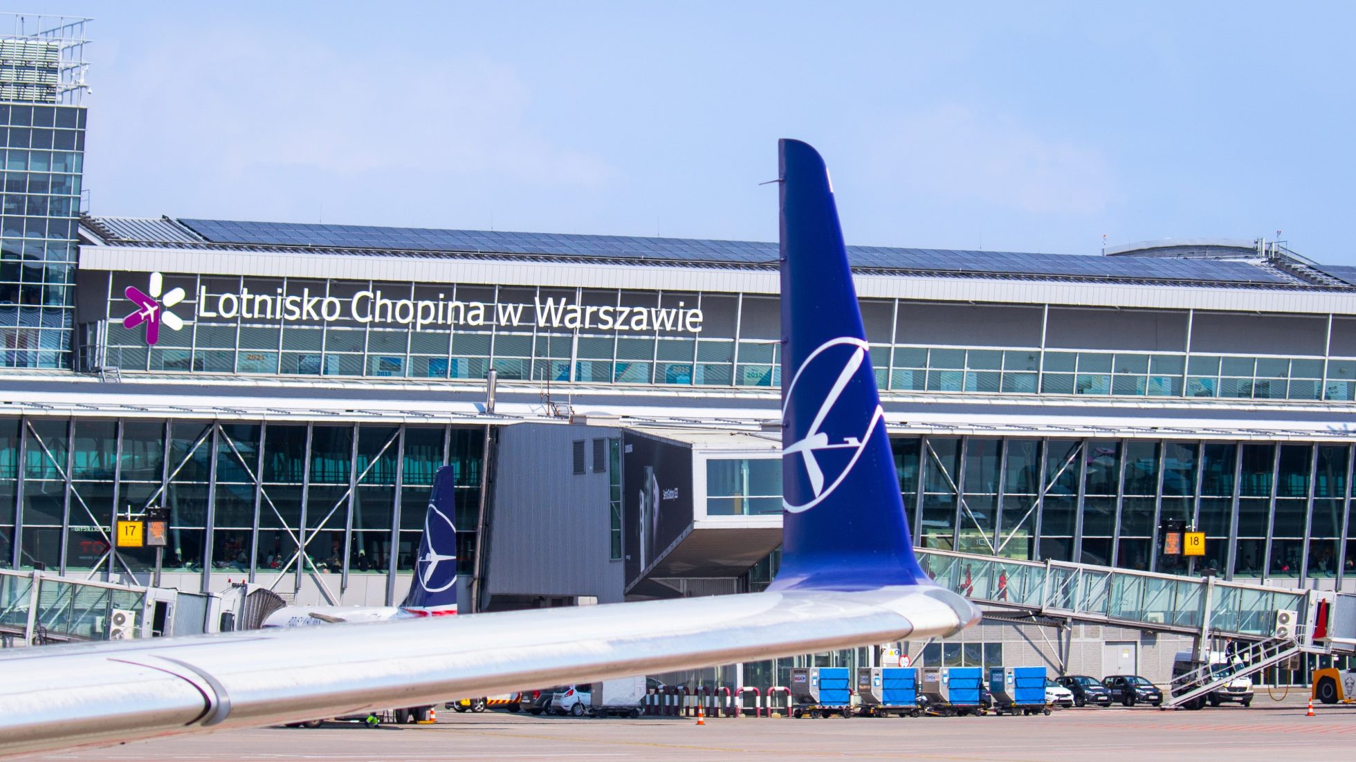 Flügel der Embraer 175 mit LOT-Kranich-Logo auf dem Winglet mit dem Terminal des Chopin-Flughafens im Hintergrund an einem sonnigen Tag