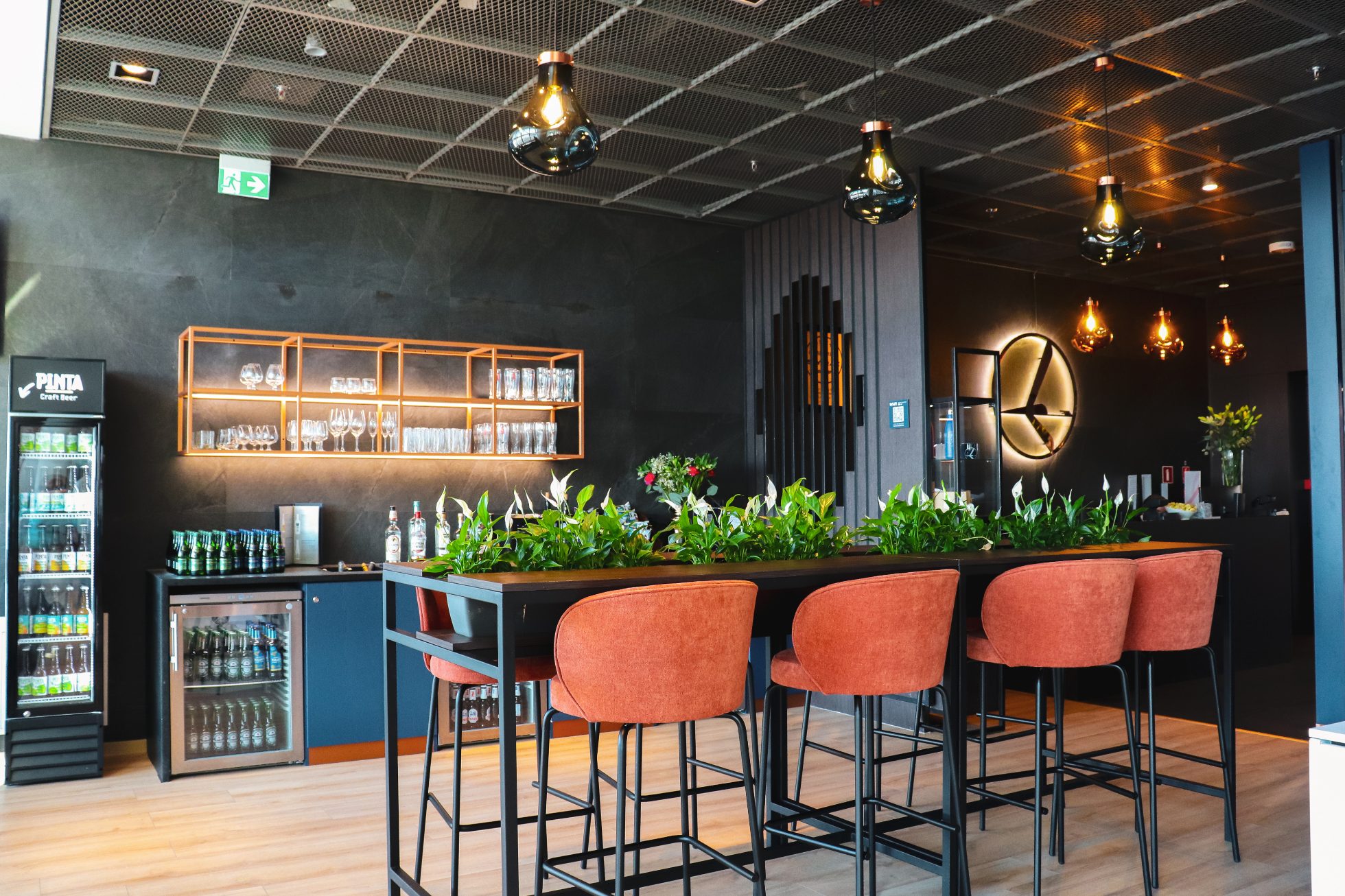 The interior of the LOT Business Lounge Mazurek – a long wooden table, orange armchairs, and an illuminated crane logo. A bar with drinks in the background.