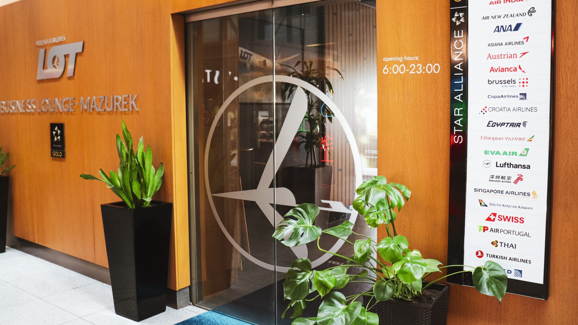 Entrance to LOT Business Lounge Mazurek - glass door with crane logo, plaque with Star Alliance member airlines, green flower in pot and silver LOT Business Lounge Mazurek inscription on wooden wall