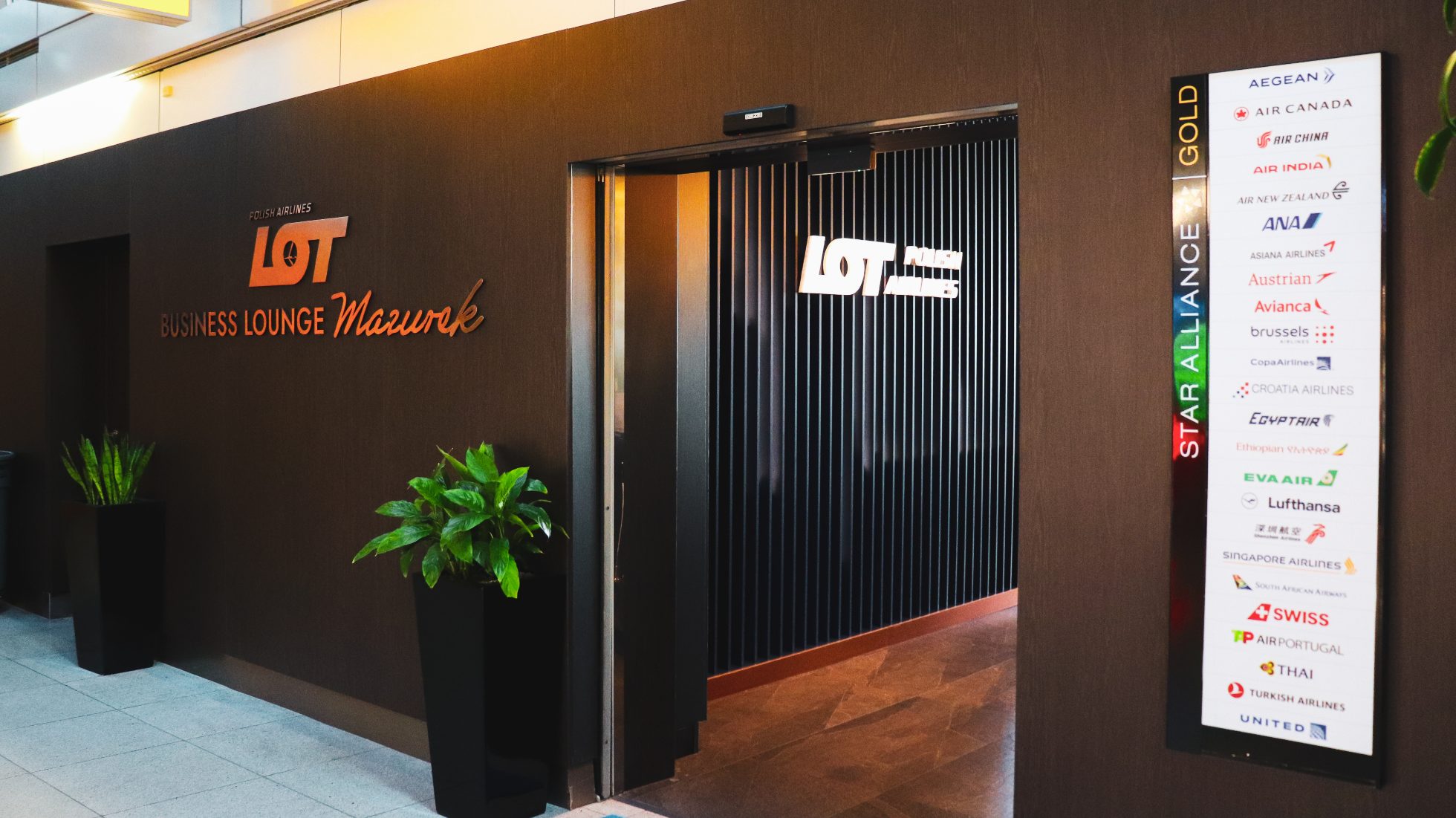 entrance to the LOT Business Lounge Mazurek – glass doors with the crane logo, a board displaying Star Alliance member airlines, a green flower in a pot, and silver lettering reading “LOT Business Lounge Mazurek” on a wooden wall
