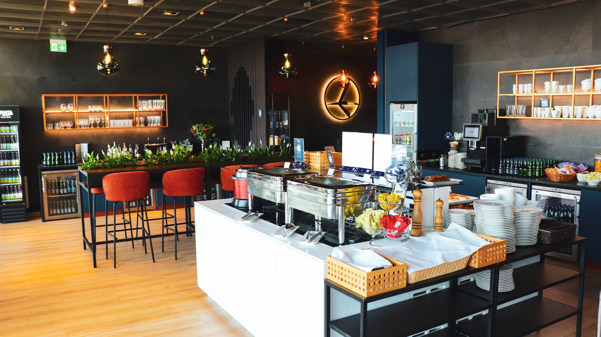 Interior of the LOT Business Lounge Maurek – dining area, long table with red armchairs, bar with snacks and drinks. Illuminated crane logo in the background.