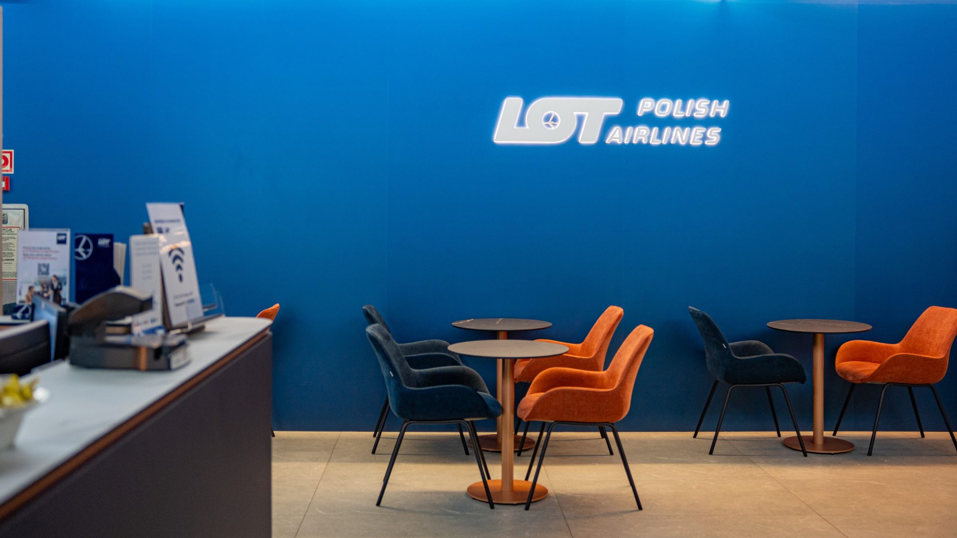 Elegancko urządzona przestrzeń - szare i pomarańczowe krzesła, granatowa ściana z logo LOT w lotniskowym salonie biznesowym Polonez