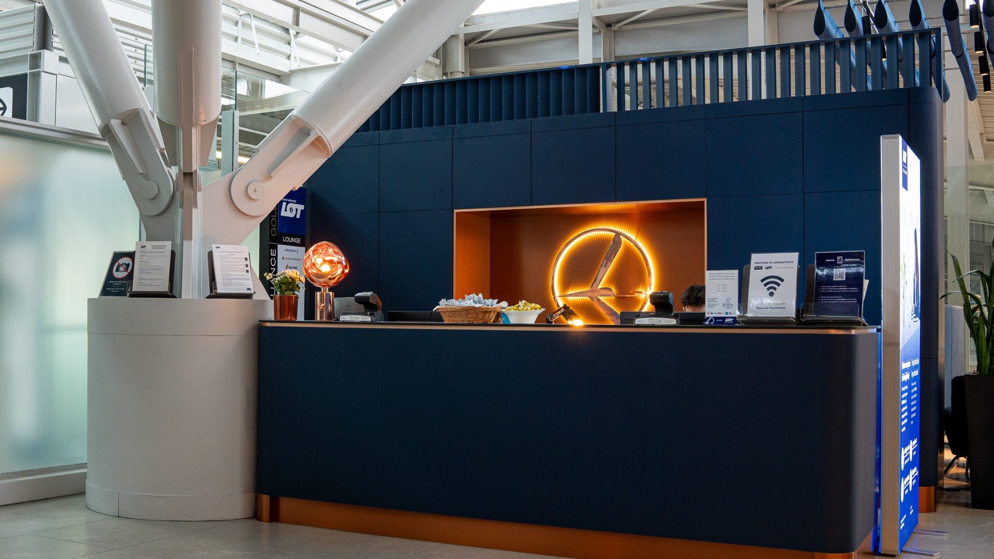 Rezeption der Polonez Business Lounge in Warschau – goldenes LOT-Neonlogo an marineblauer Wand, Snacks für Gäste auf der Theke