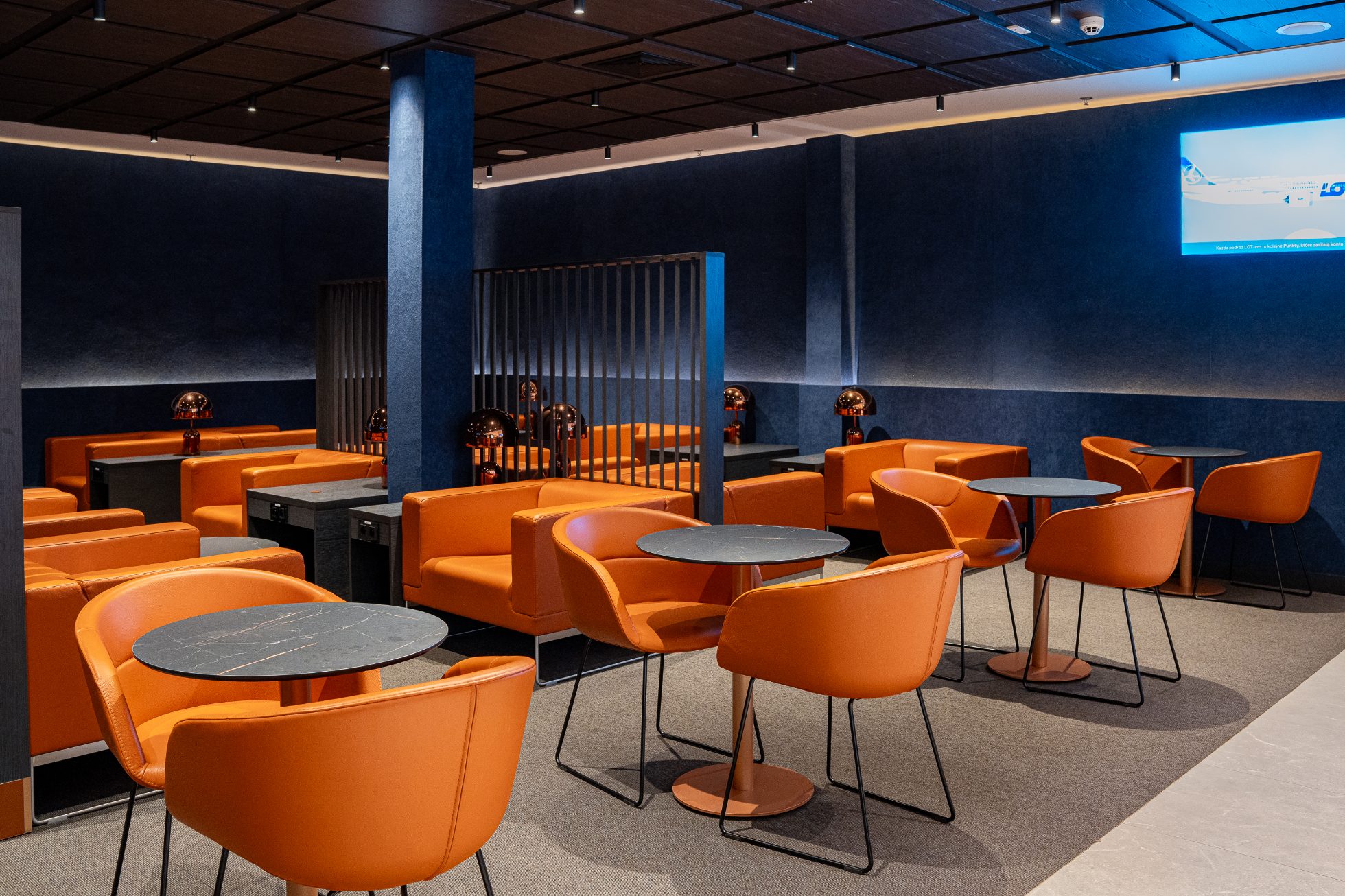 Polonez Gold Lounge - an open space with orange chairs and sofas, navy blue walls with the Crane logo. Catering area with hot meals, snacks and drinks. Relaxation area with armchairs and dimmed lighting.