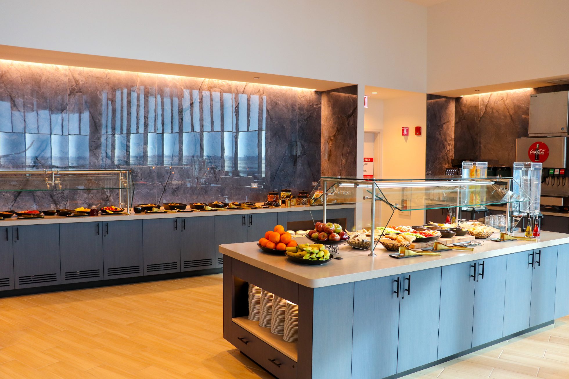LOT Business Lounge in Chicago - reception area with illuminated LOT logo. Public space with yellow sofas, copper lamps and tables with white marble tops. Blue carpets. High ceiling and huge panoramic windows overlooking the airport. Large bar with backlights