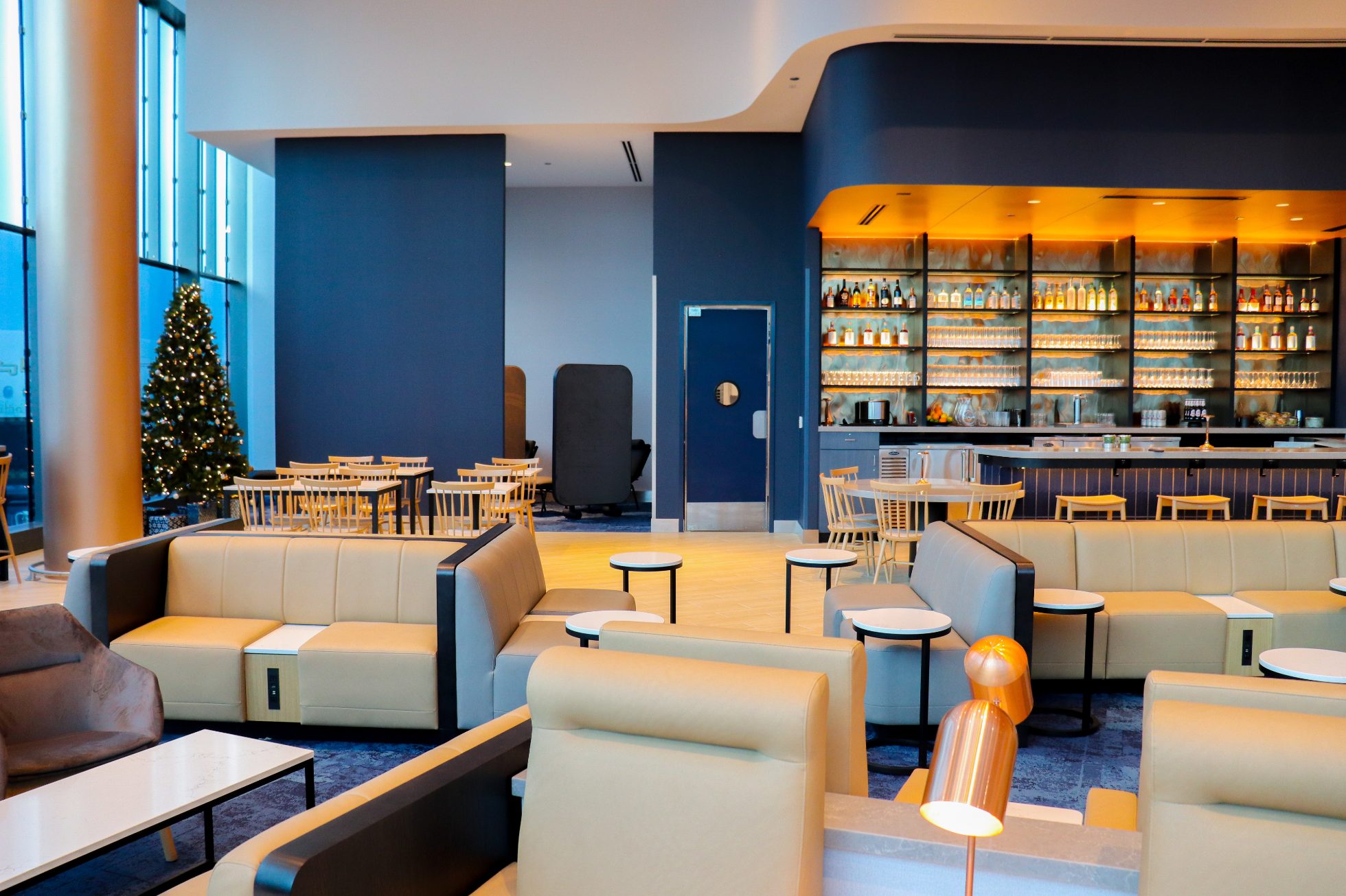 LOT Business Lounge in Chicago - reception area with illuminated LOT logo. Public space with yellow sofas, copper lamps and tables with white marble tops. Blue carpets. High ceiling and huge panoramic windows overlooking the airport. Large bar with backlights