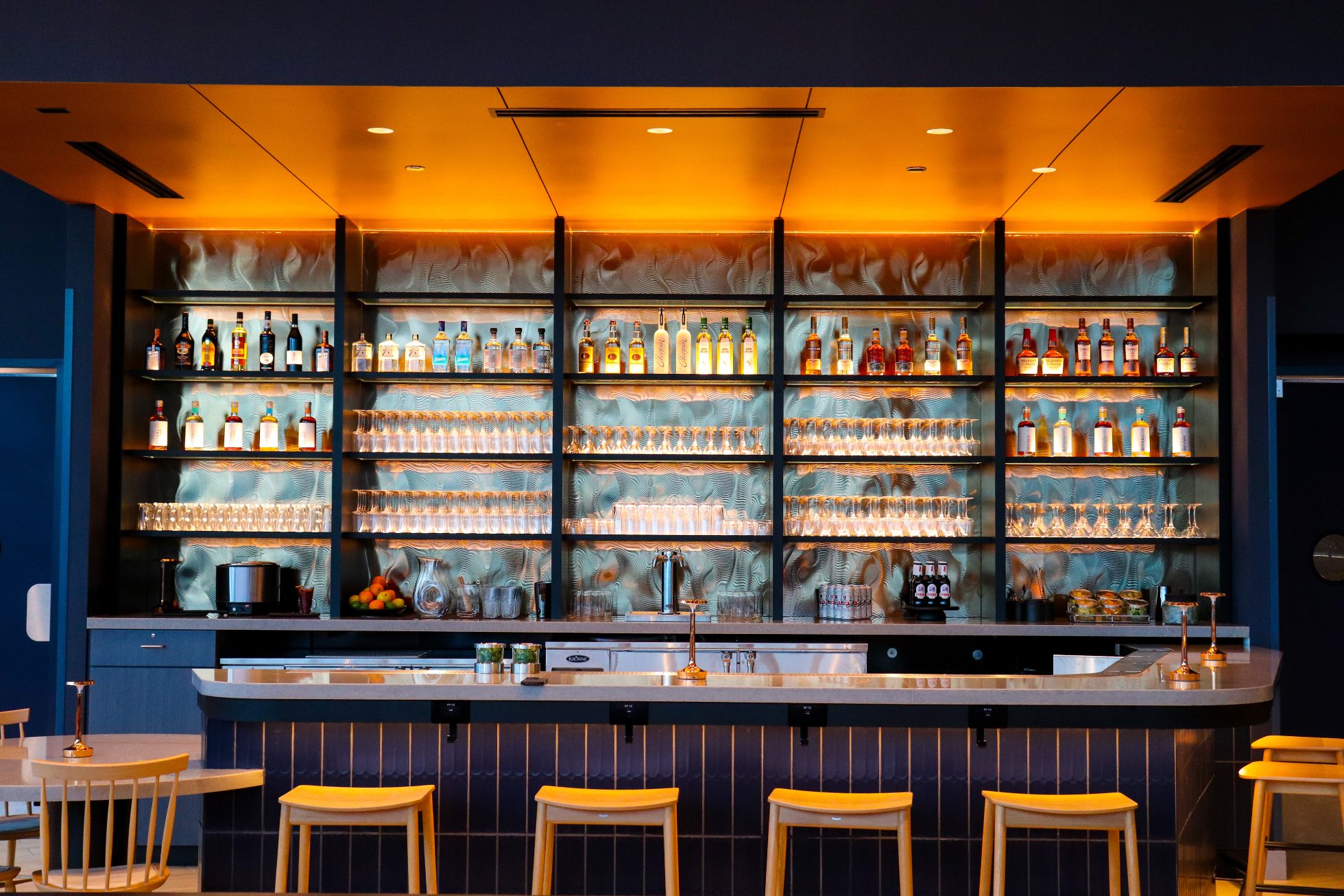 LOT Business Lounge in Chicago - reception area with illuminated LOT logo. Public space with yellow sofas, copper lamps and tables with white marble tops. Blue carpets. High ceiling and huge panoramic windows overlooking the airport. Large bar with backlights