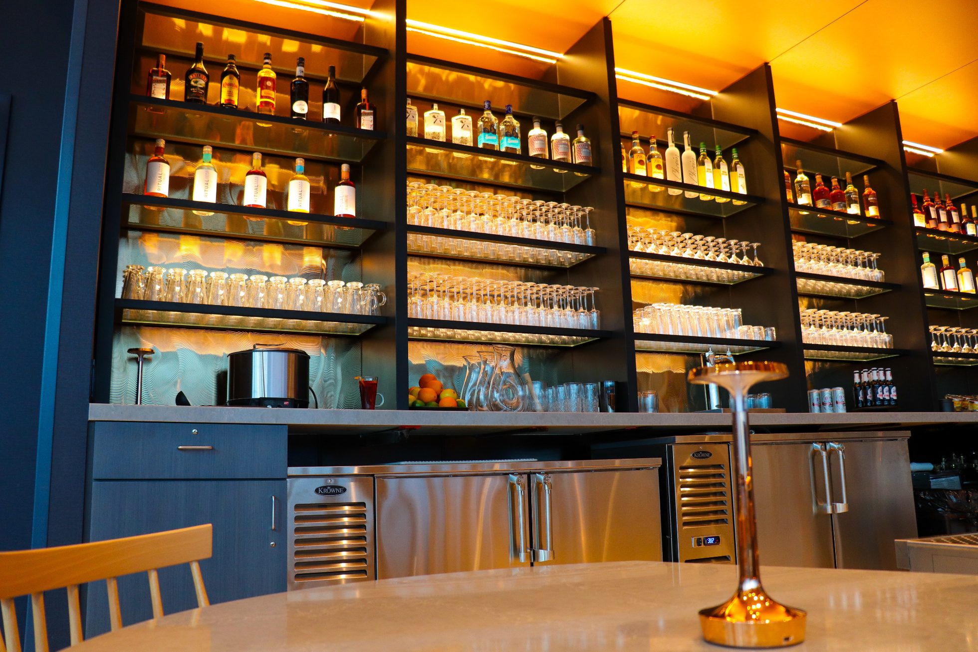 LOT Business Lounge in Chicago - reception area with illuminated LOT logo. Public space with yellow sofas, copper lamps and tables with white marble tops. Blue carpets. High ceiling and huge panoramic windows overlooking the airport. Large bar with backlights