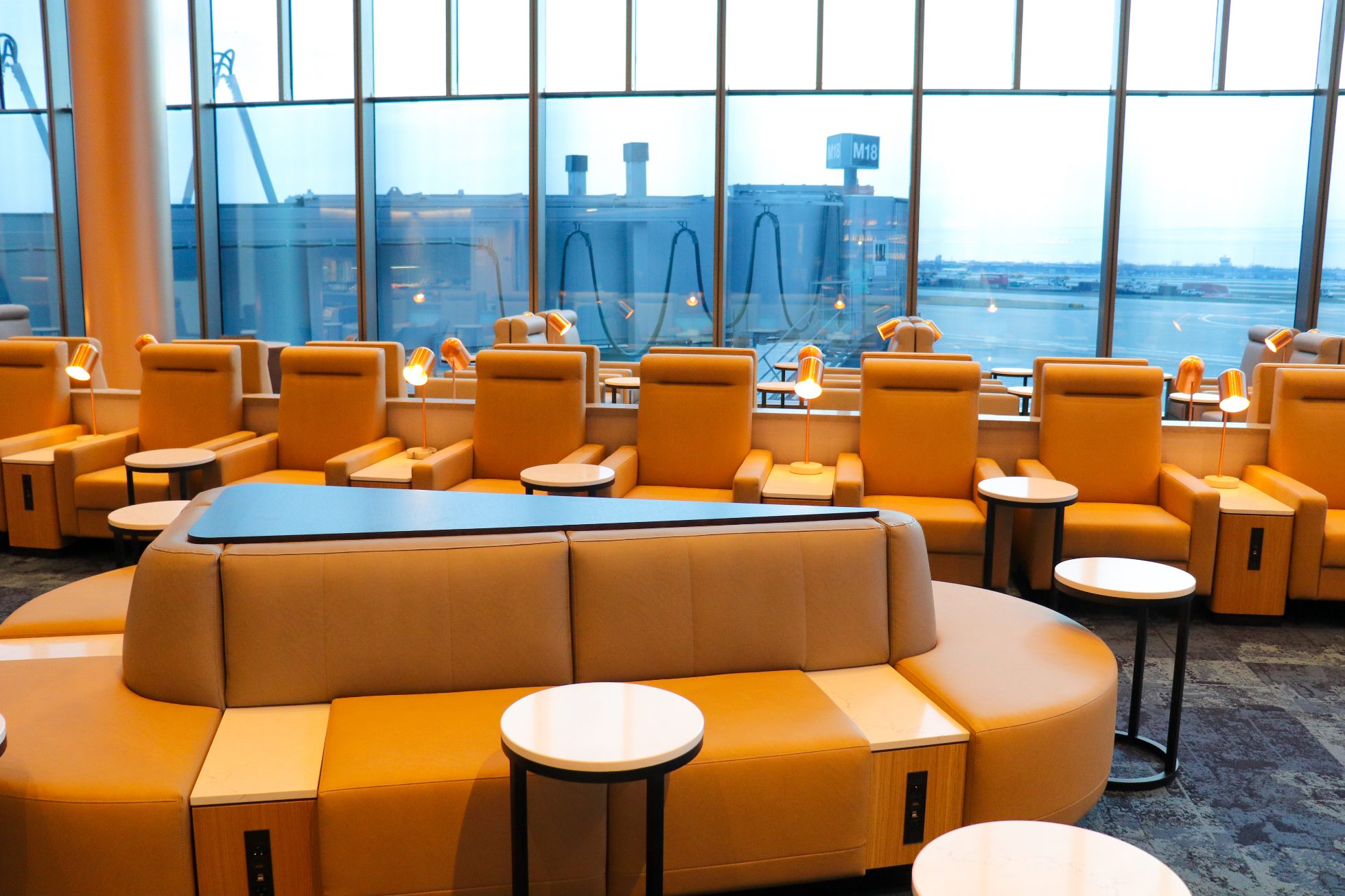 LOT Business Lounge in Chicago - reception area with illuminated LOT logo. Public space with yellow sofas, copper lamps and tables with white marble tops. Blue carpets. High ceiling and huge panoramic windows overlooking the airport. Large bar with backlights