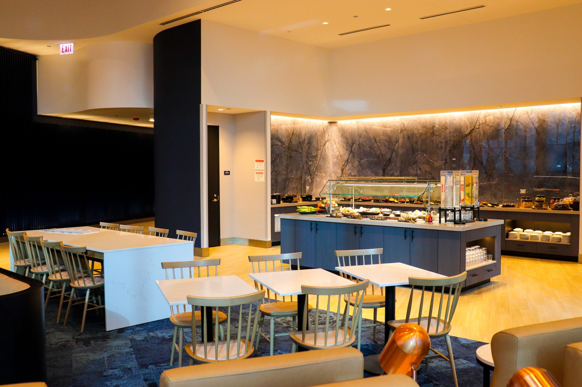 LOT Business Lounge in Chicago - reception area with illuminated LOT logo. Public space with yellow sofas, copper lamps and tables with white marble tops. Blue carpets. High ceiling and huge panoramic windows overlooking the airport. Large bar with backlights