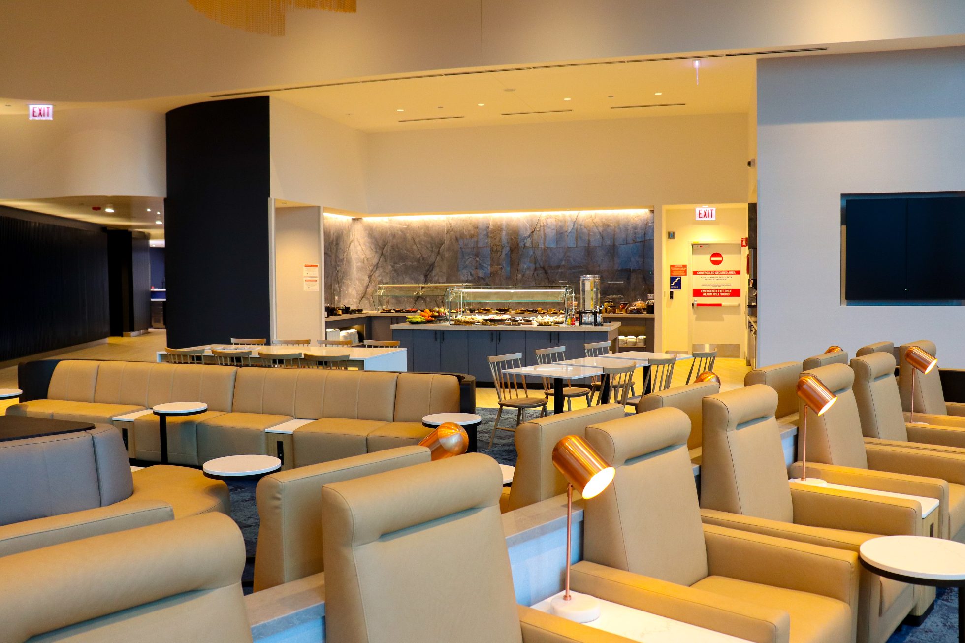 LOT Business Lounge in Chicago - reception area with illuminated LOT logo. Public space with yellow sofas, copper lamps and tables with white marble tops. Blue carpets. High ceiling and huge panoramic windows overlooking the airport. Large bar with backlights