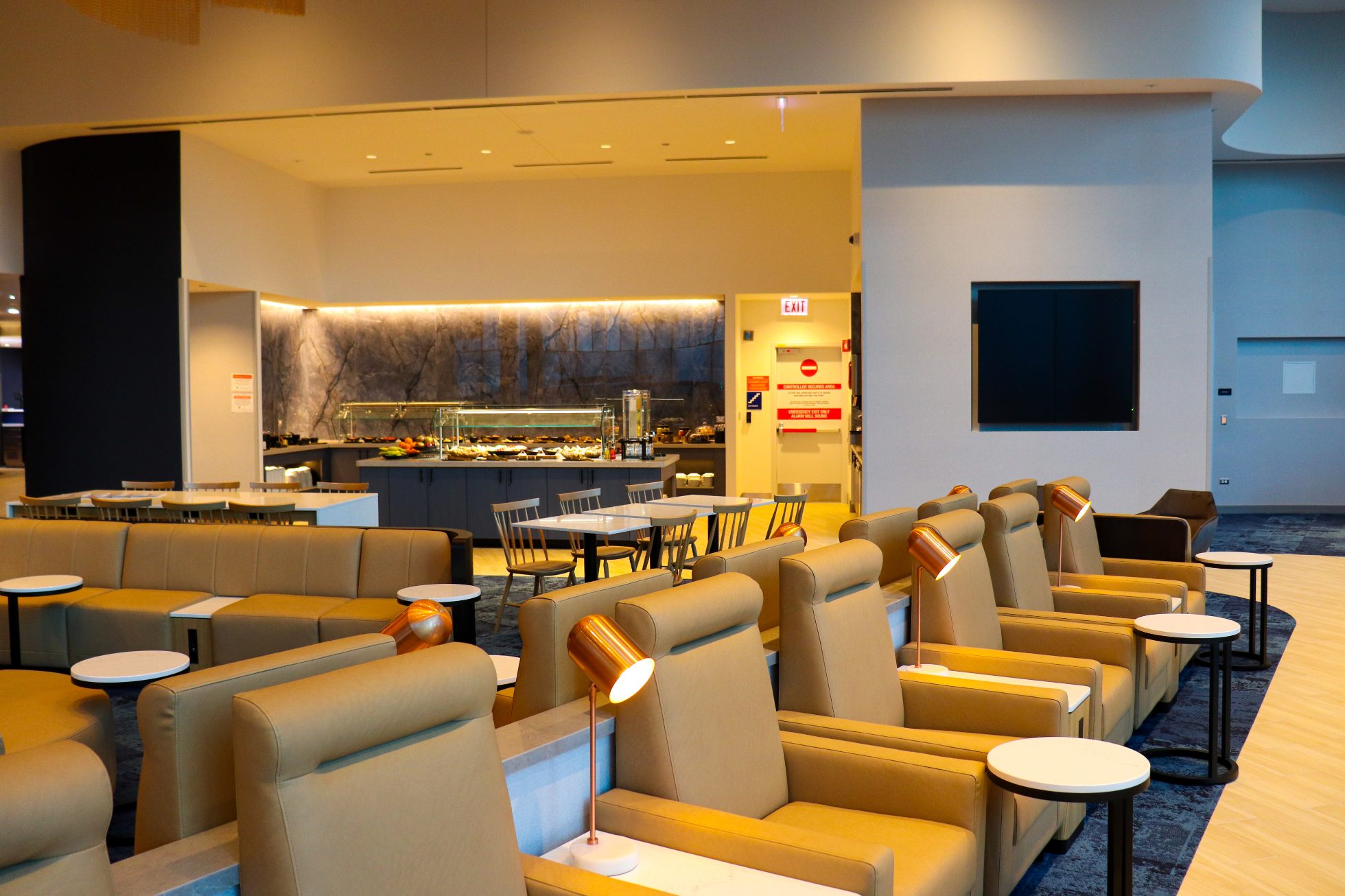 LOT Business Lounge in Chicago - reception area with illuminated LOT logo. Public space with yellow sofas, copper lamps and tables with white marble tops. Blue carpets. High ceiling and huge panoramic windows overlooking the airport. Large bar with backlights