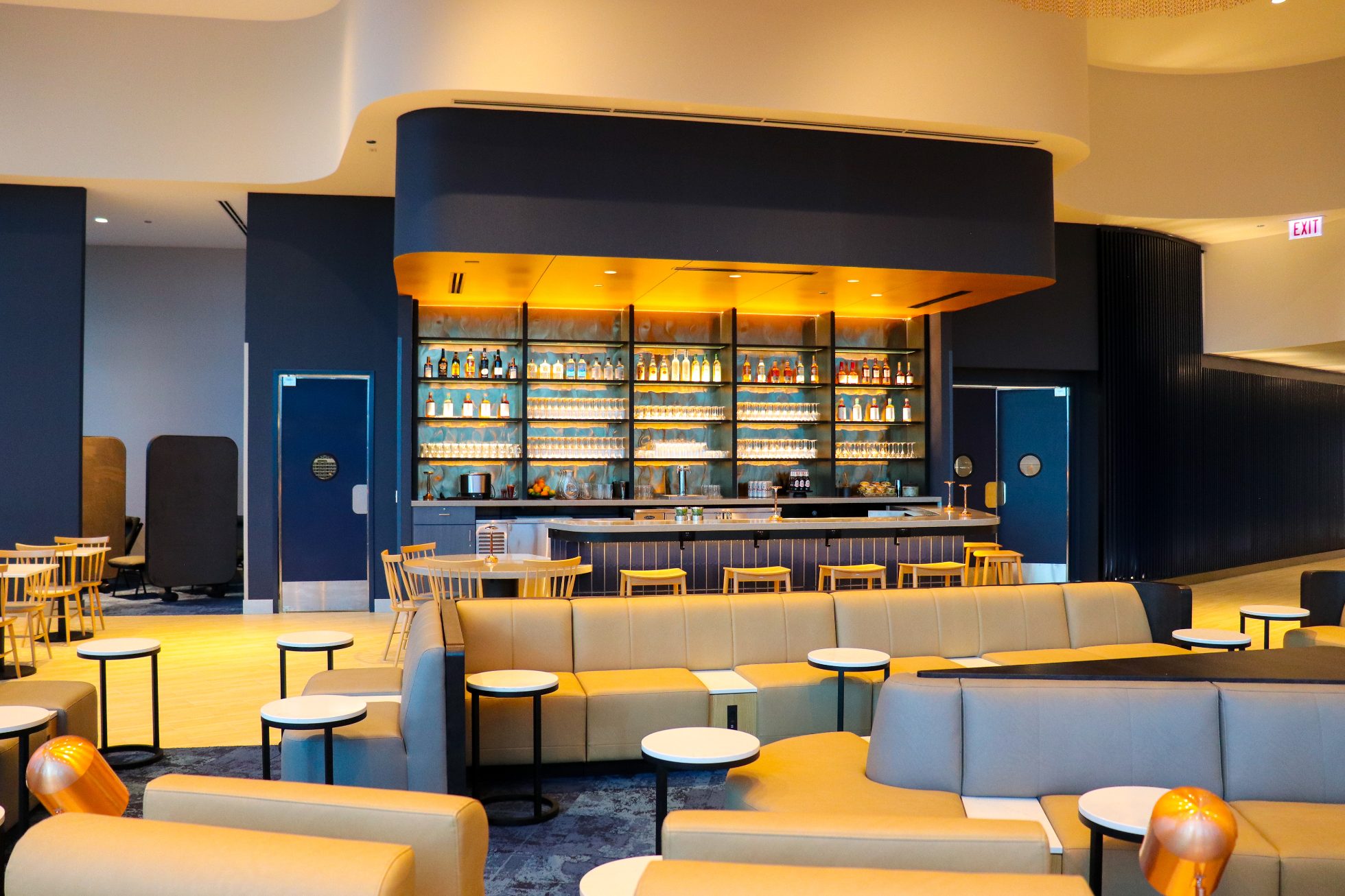 LOT Business Lounge in Chicago - reception area with illuminated LOT logo. Public space with yellow sofas, copper lamps and tables with white marble tops. Blue carpets. High ceiling and huge panoramic windows overlooking the airport. Large bar with backlights