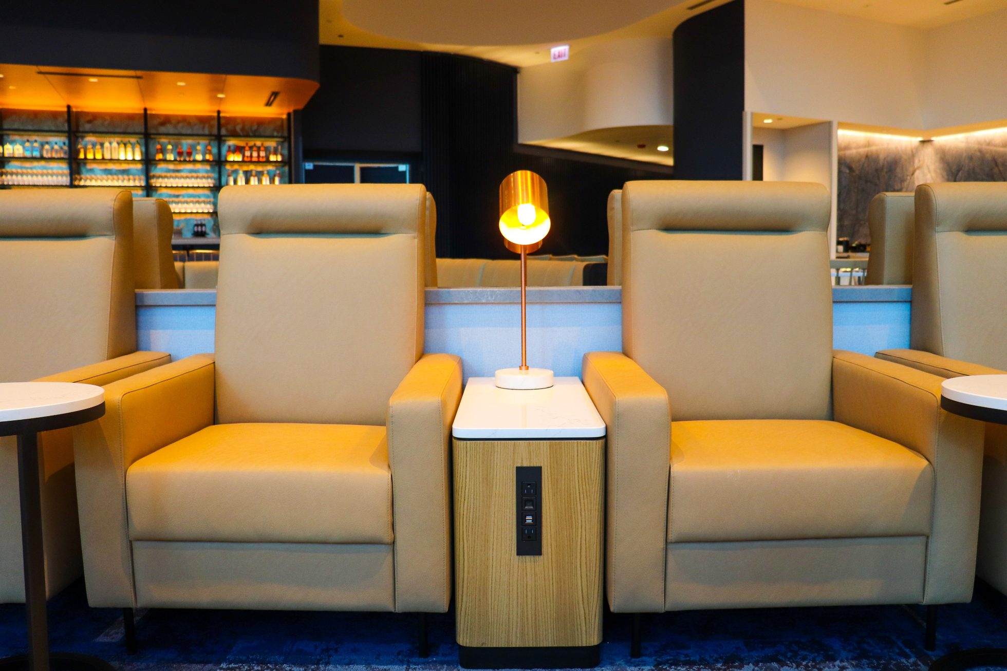 LOT Business Lounge in Chicago - reception area with illuminated LOT logo. Public space with yellow sofas, copper lamps and tables with white marble tops. Blue carpets. High ceiling and huge panoramic windows overlooking the airport. Large bar with backlights