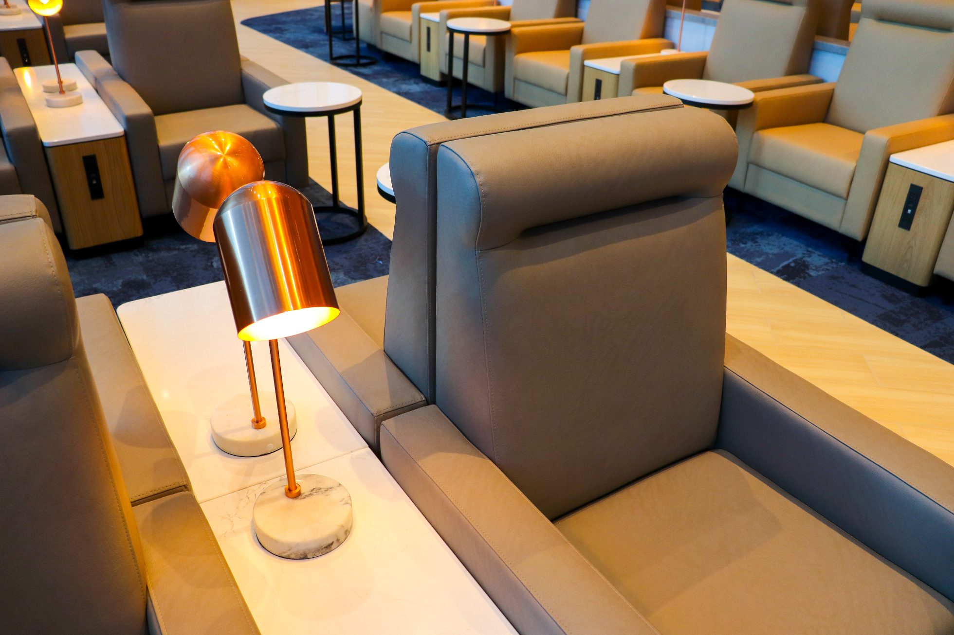 LOT Business Lounge in Chicago - reception area with illuminated LOT logo. Public space with yellow sofas, copper lamps and tables with white marble tops. Blue carpets. High ceiling and huge panoramic windows overlooking the airport. Large bar with backlights