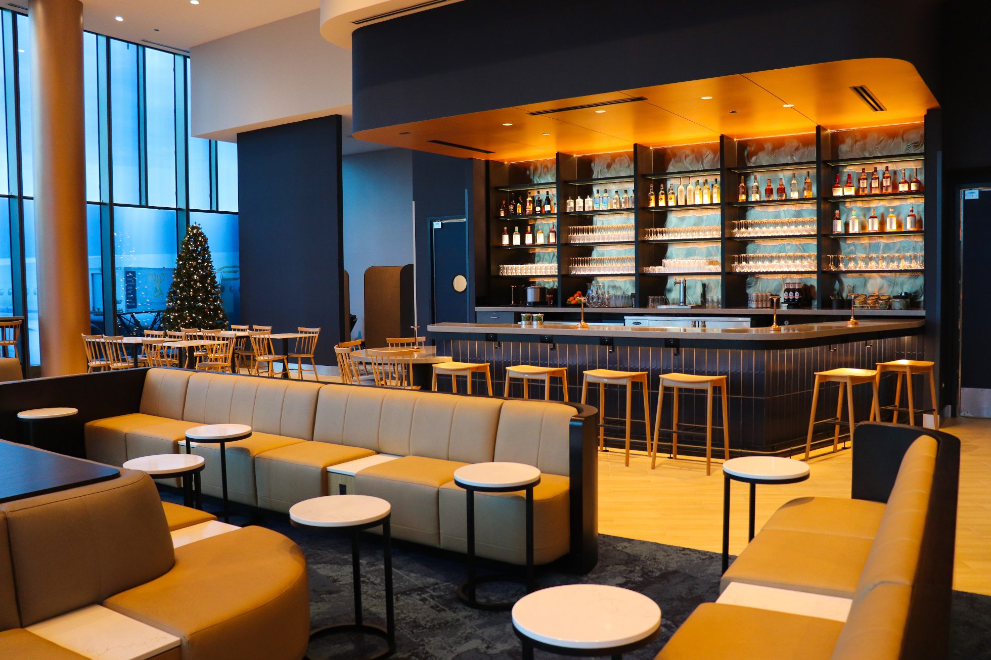 LOT Business Lounge in Chicago - reception area with illuminated LOT logo. Public space with yellow sofas, copper lamps and tables with white marble tops. Blue carpets. High ceiling and huge panoramic windows overlooking the airport. Large bar with backlights