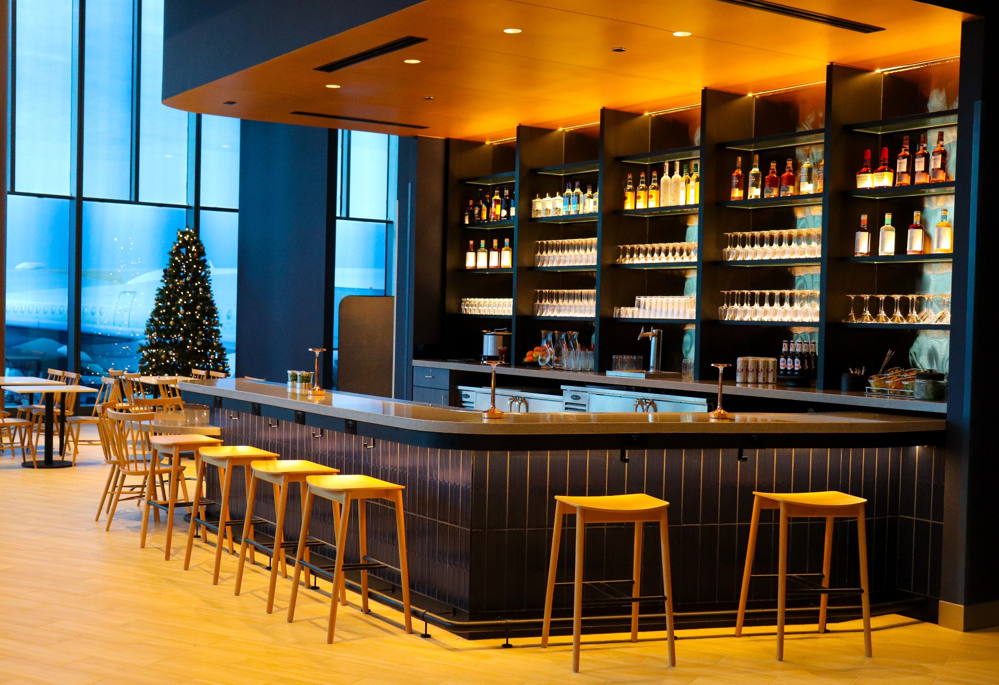 LOT Business Lounge in Chicago - reception area with illuminated LOT logo. Public space with yellow sofas, copper lamps and tables with white marble tops. Blue carpets. High ceiling and huge panoramic windows overlooking the airport. Large bar with backlights