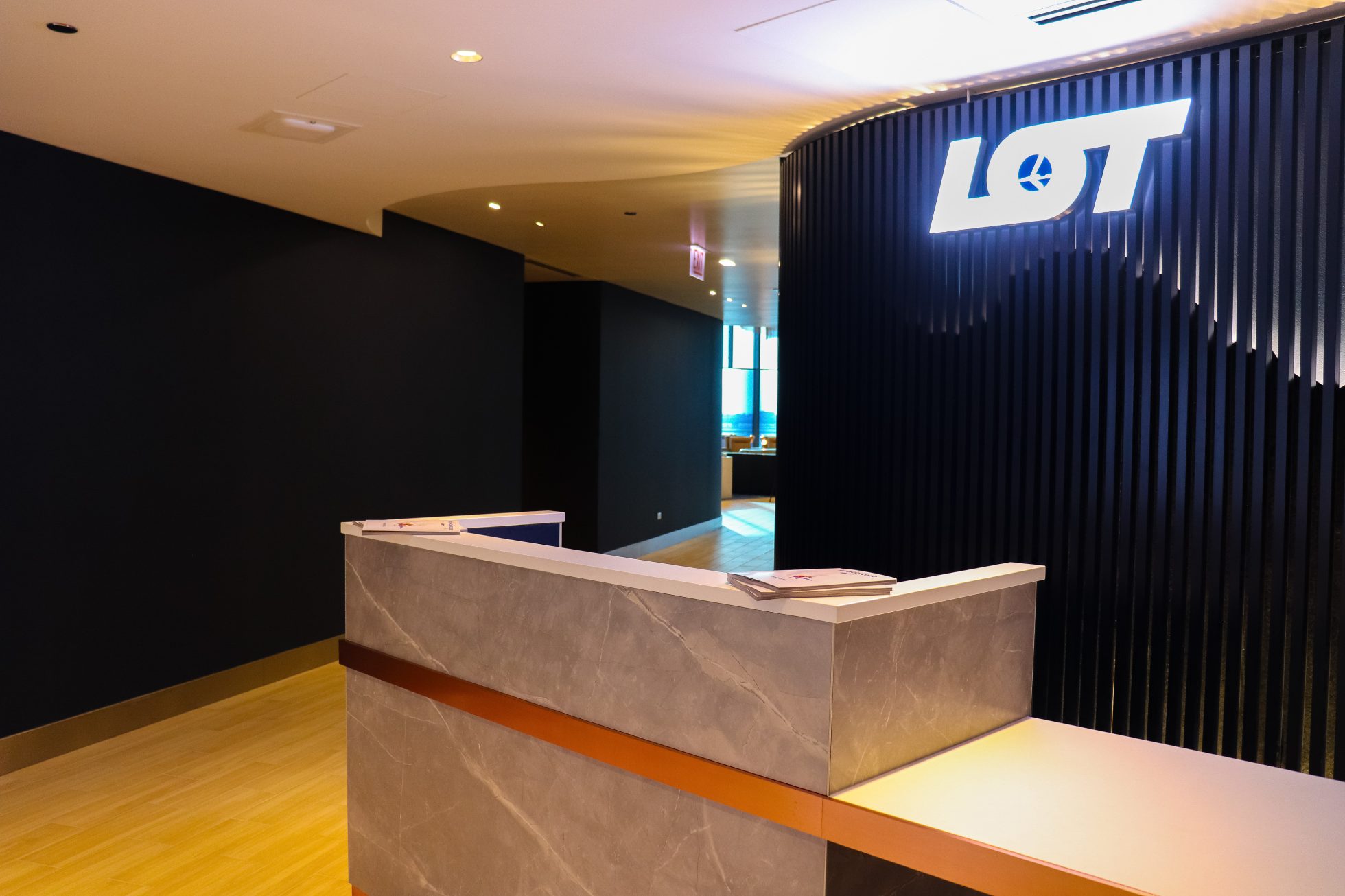 LOT Business Lounge in Chicago - reception area with illuminated LOT logo. Public space with yellow sofas, copper lamps and tables with white marble tops. Blue carpets. High ceiling and huge panoramic windows overlooking the airport. Large bar with backlights