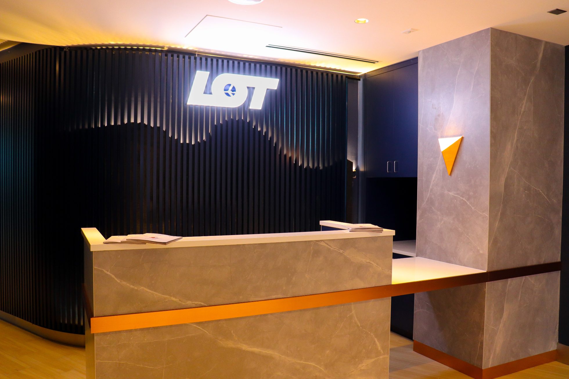 LOT Business Lounge in Chicago - reception area with illuminated LOT logo. Public space with yellow sofas, copper lamps and tables with white marble tops. Blue carpets. High ceiling and huge panoramic windows overlooking the airport. Large bar with backlights