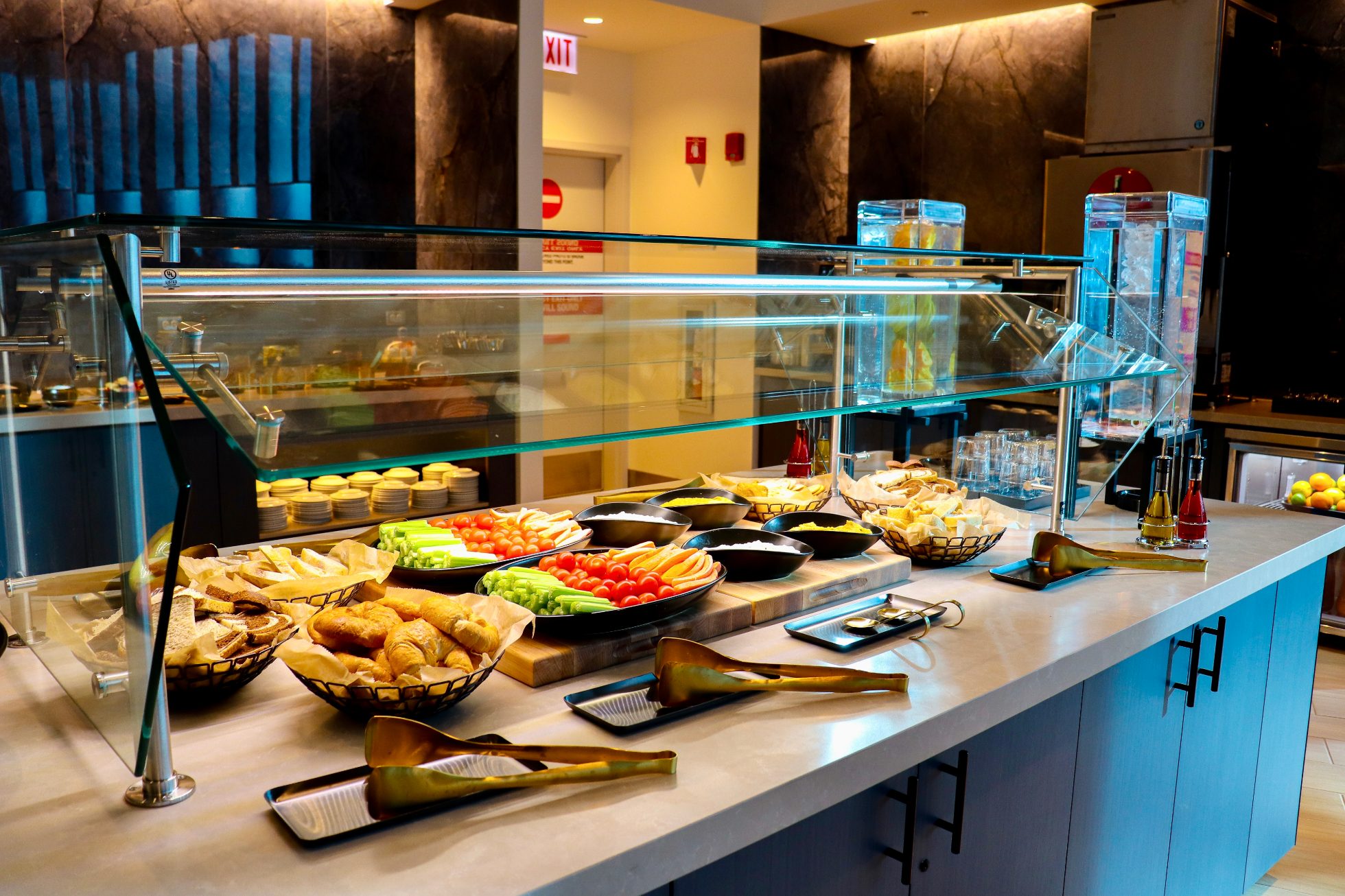 LOT Business Lounge in Chicago - reception area with illuminated LOT logo. Public space with yellow sofas, copper lamps and tables with white marble tops. Blue carpets. High ceiling and huge panoramic windows overlooking the airport. Large bar with backlights