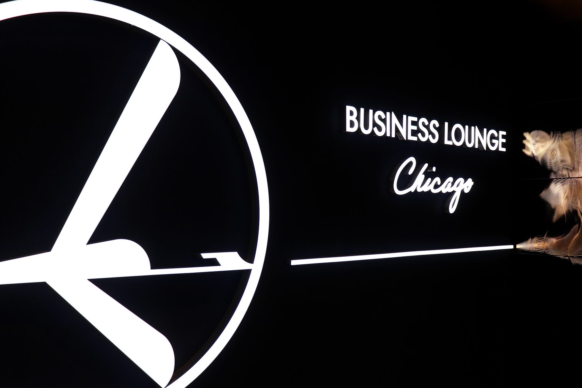 LOT Business Lounge in Chicago - reception area with illuminated LOT logo. Public space with yellow sofas, copper lamps and tables with white marble tops. Blue carpets. High ceiling and huge panoramic windows overlooking the airport. Large bar with backlights