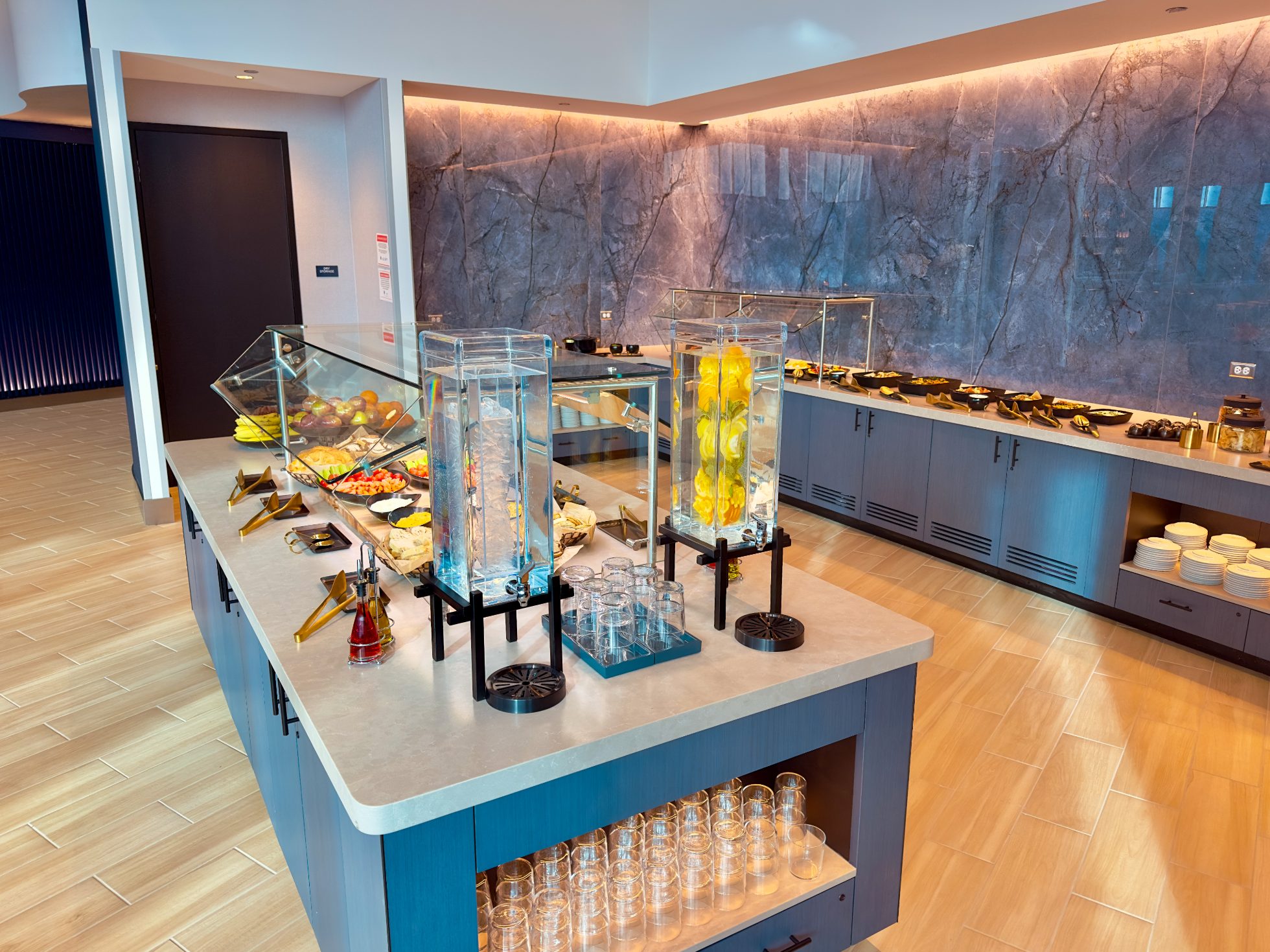 LOT Business Lounge in Chicago - reception area with illuminated LOT logo. Public space with yellow sofas, copper lamps and tables with white marble tops. Blue carpets. High ceiling and huge panoramic windows overlooking the airport. Large bar with backlights