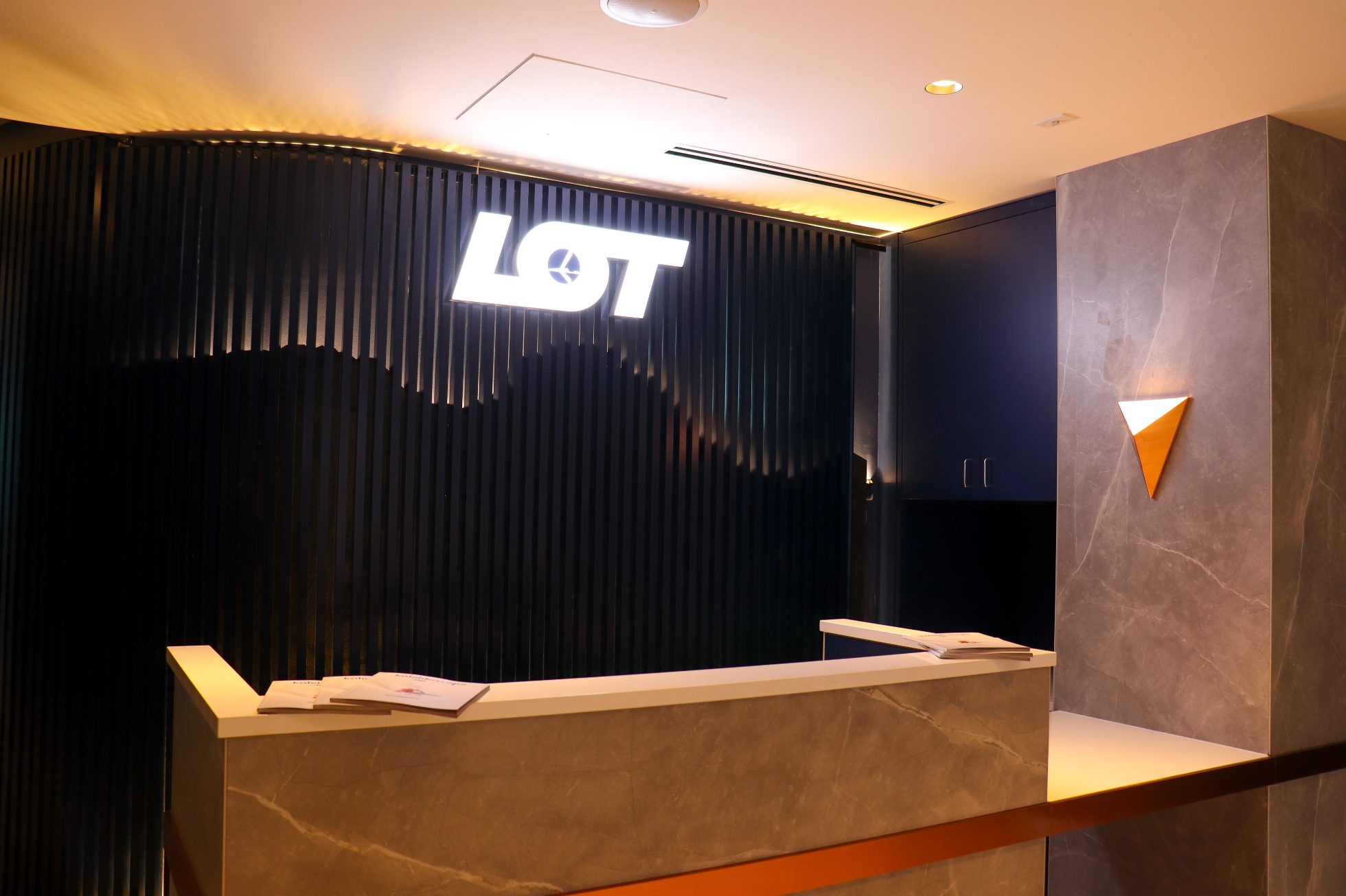 LOT Business Lounge in Chicago - reception area with illuminated LOT logo. Public space with yellow sofas, copper lamps and tables with white marble tops. Blue carpets. High ceiling and huge panoramic windows overlooking the airport. Large bar with backlights