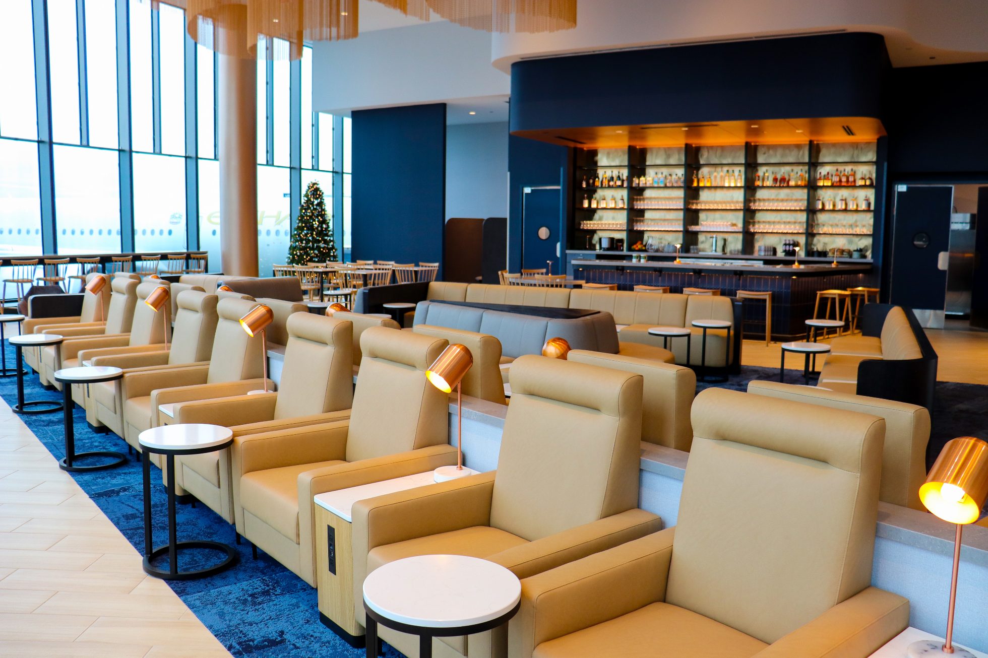 LOT Business Lounge in Chicago - reception area with illuminated LOT logo. Public space with yellow sofas, copper lamps and tables with white marble tops. Blue carpets. High ceiling and huge panoramic windows overlooking the airport. Large bar with backlights