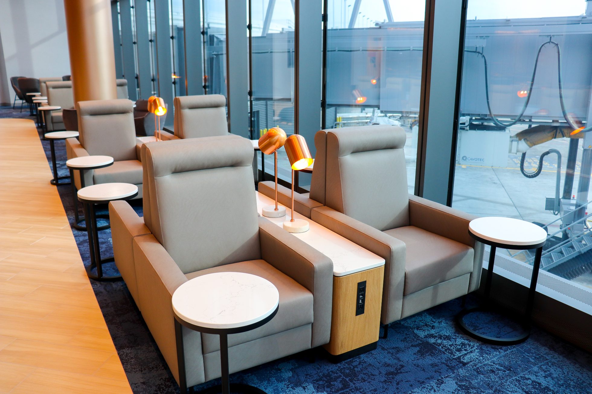 LOT Business Lounge in Chicago - reception area with illuminated LOT logo. Public space with yellow sofas, copper lamps and tables with white marble tops. Blue carpets. High ceiling and huge panoramic windows overlooking the airport. Large bar with backlights