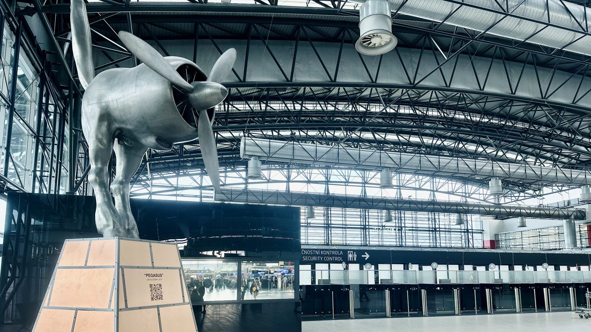 Terminal lotniska w Pradze. W tle widoczne są tablice informacyjne i sklepy duty-free.