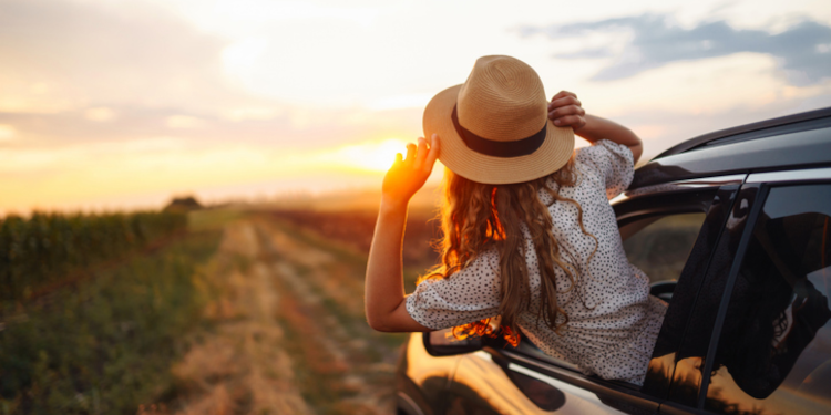 A woman leaning out of the front window of a car at sunset among fields.