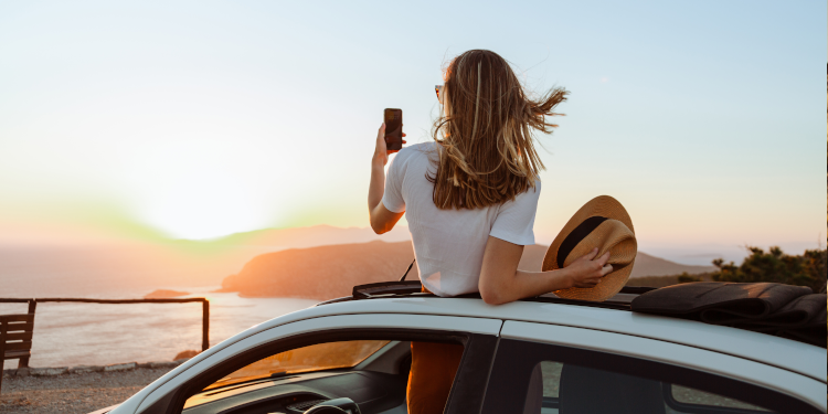 A woman taking a picture of the sunset over the sea from a car.