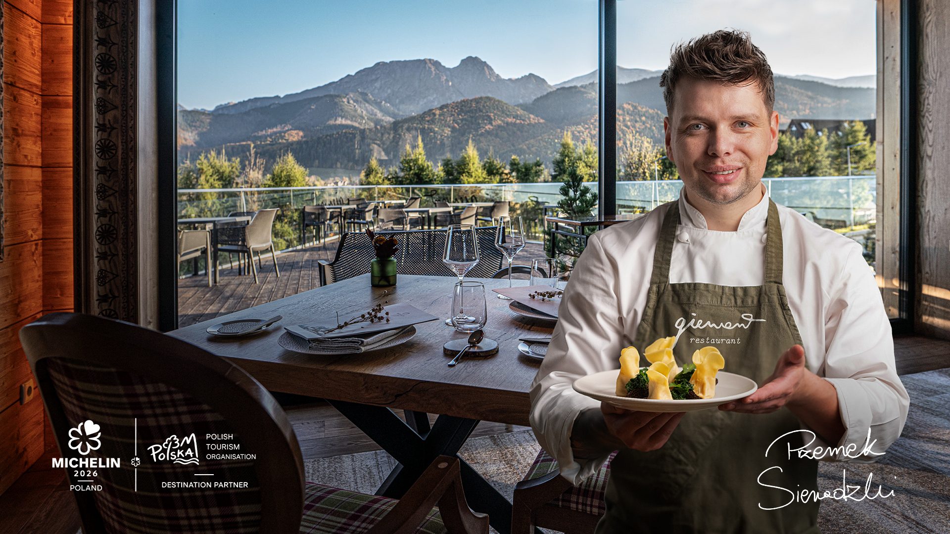 Grafika z Szefem Kuchni Przemkiem Sieradzkim w restauracji Giewont. Szef trzyma danie - vege pierogi ze smardzami. W tle widać stolik nakryty zastawą obiadową a za oknem góra Giewont