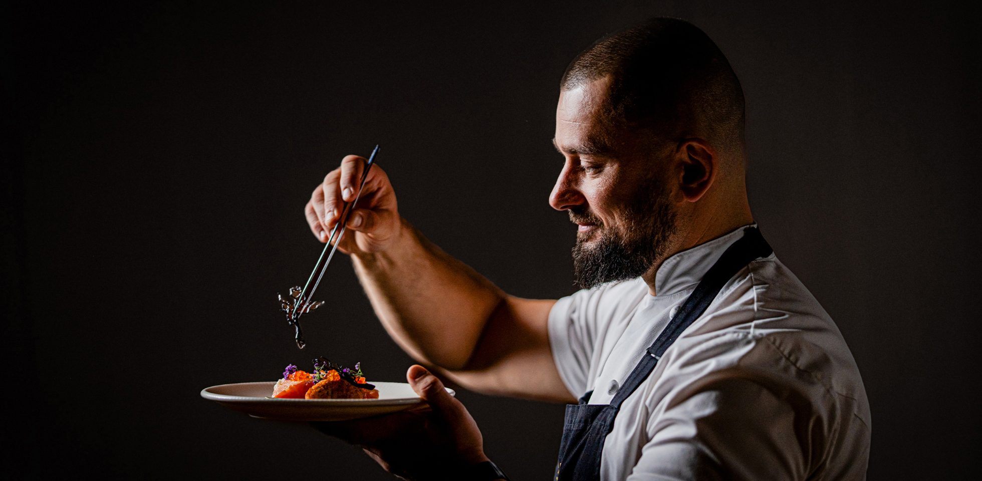 Bartosz Szymczak, head chef at Rozbrat20 restaurant, decorating his dish against a dark background.