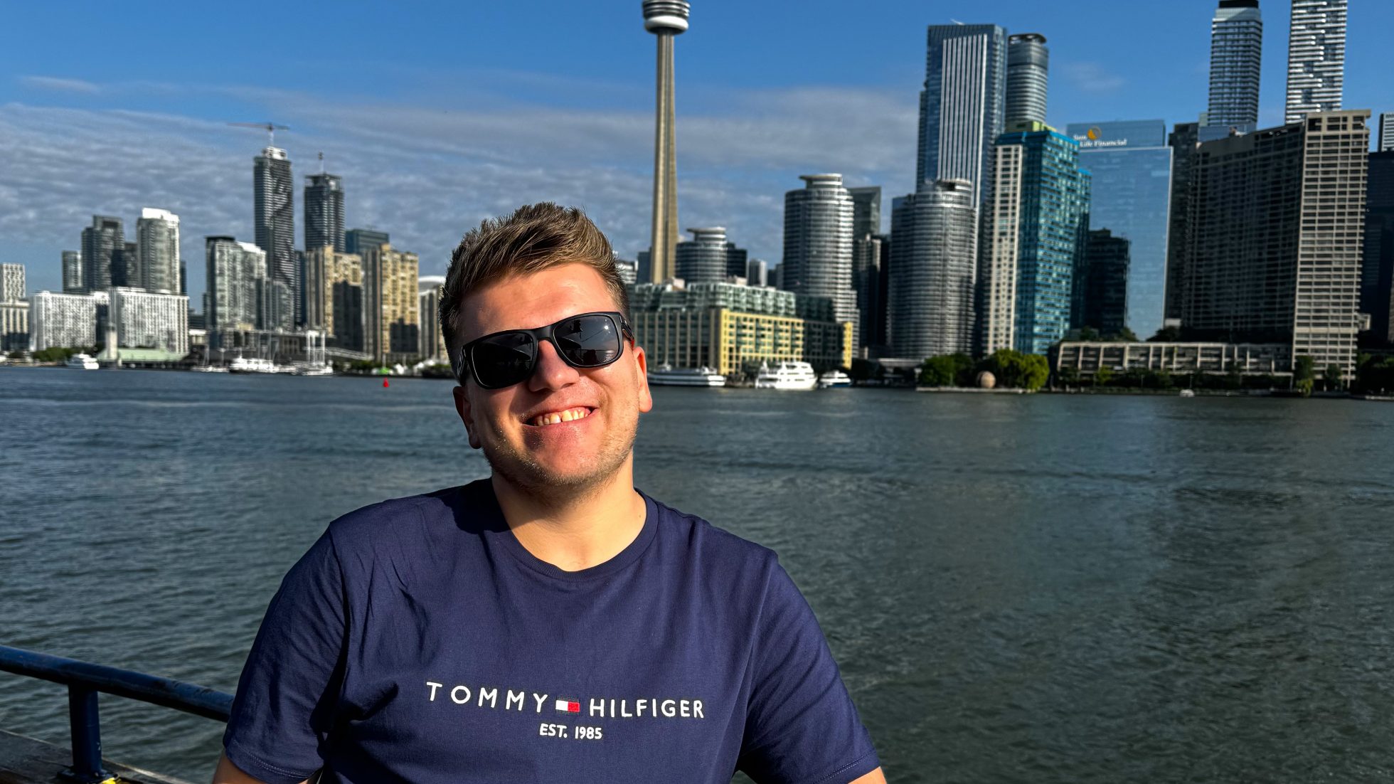 Mężczyzna w okularach przeciwsłonecznych w granatowej koszulce i panorama Toronto w tle w słoneczny dzień