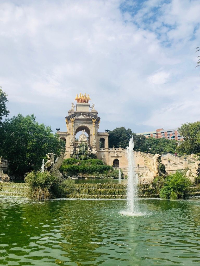 Park de la Ciutadella w Barcelonie. Ludzie spacerują po parku, w tle widać zielone drzewa i fontannę.
