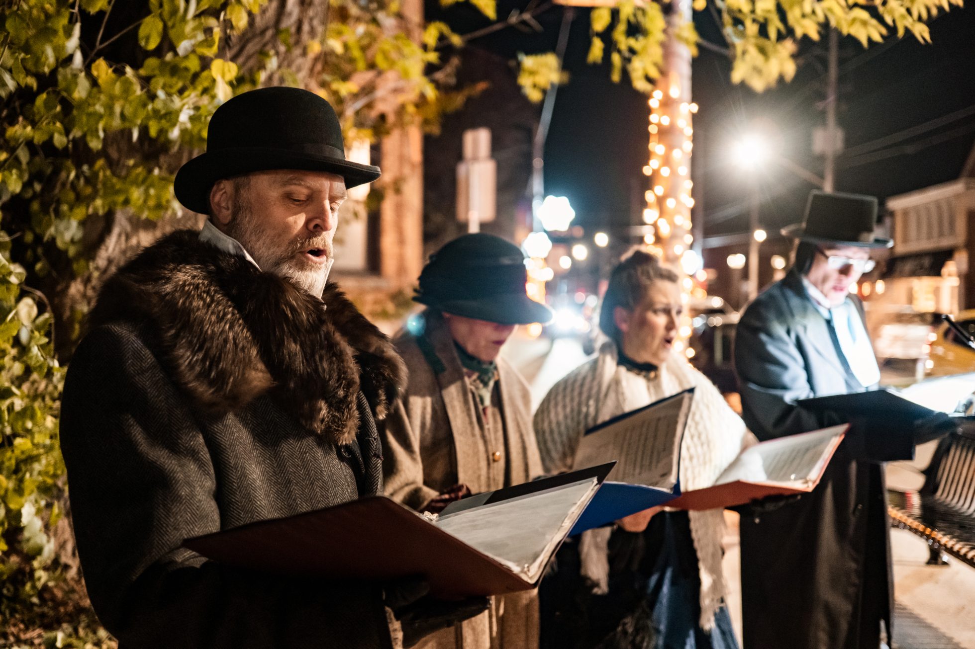 Dojrzali ludzie śpiewają kolędy na świeżym powietrzu w nocy. Ubrani są w stroje z epoki. Na zewnątrz ulicy miejskiej z wystrojem świątecznym, tradycje bożonarodzeniowe w Polsce